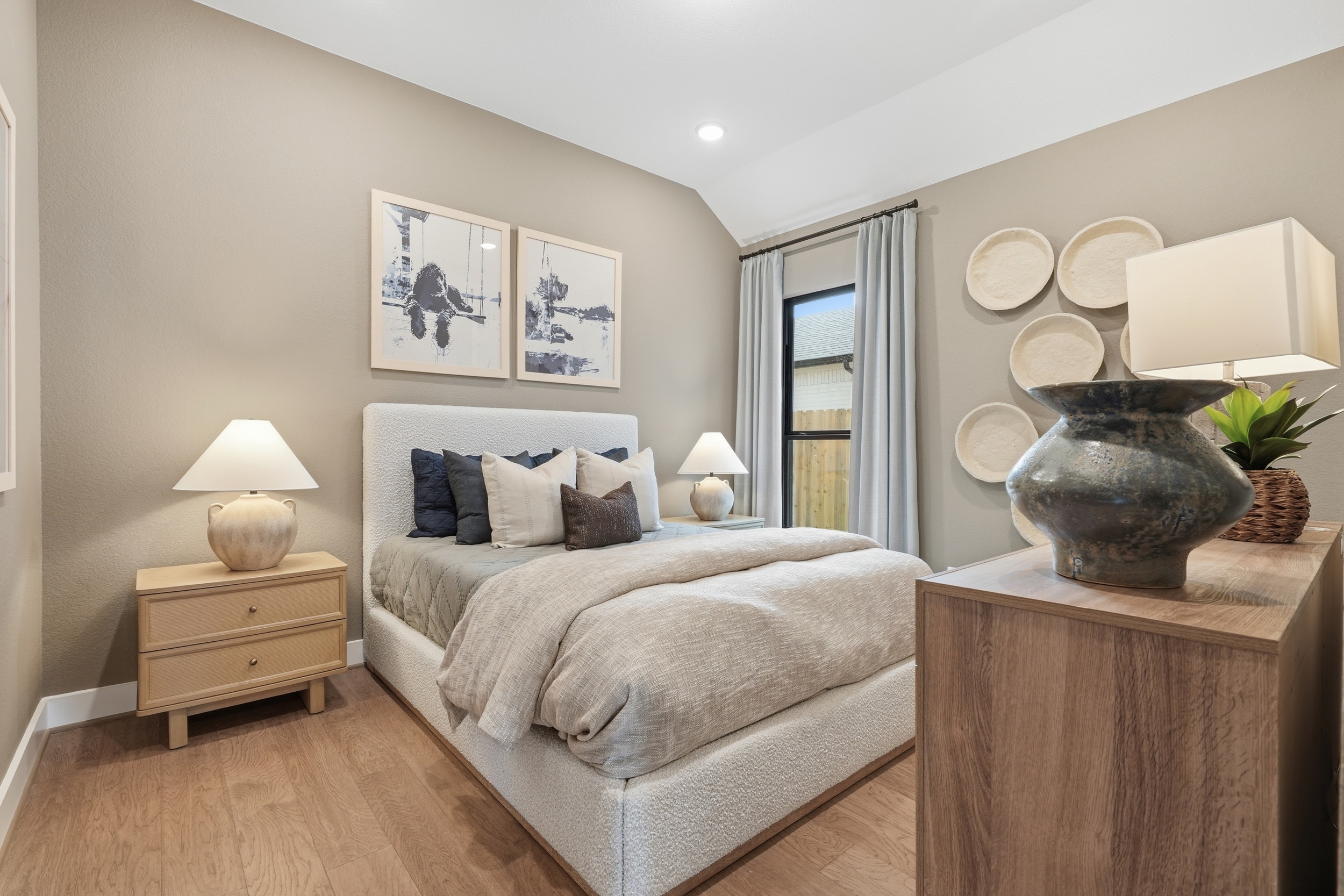 A cozy bedroom in one of the beautiful new homes in Rosharon, TX, features a neatly made bed with neutral bedding, decorative pillows, bedside tables with lamps, a dresser with a vase, wall art, and woven baskets accenting the tan walls.