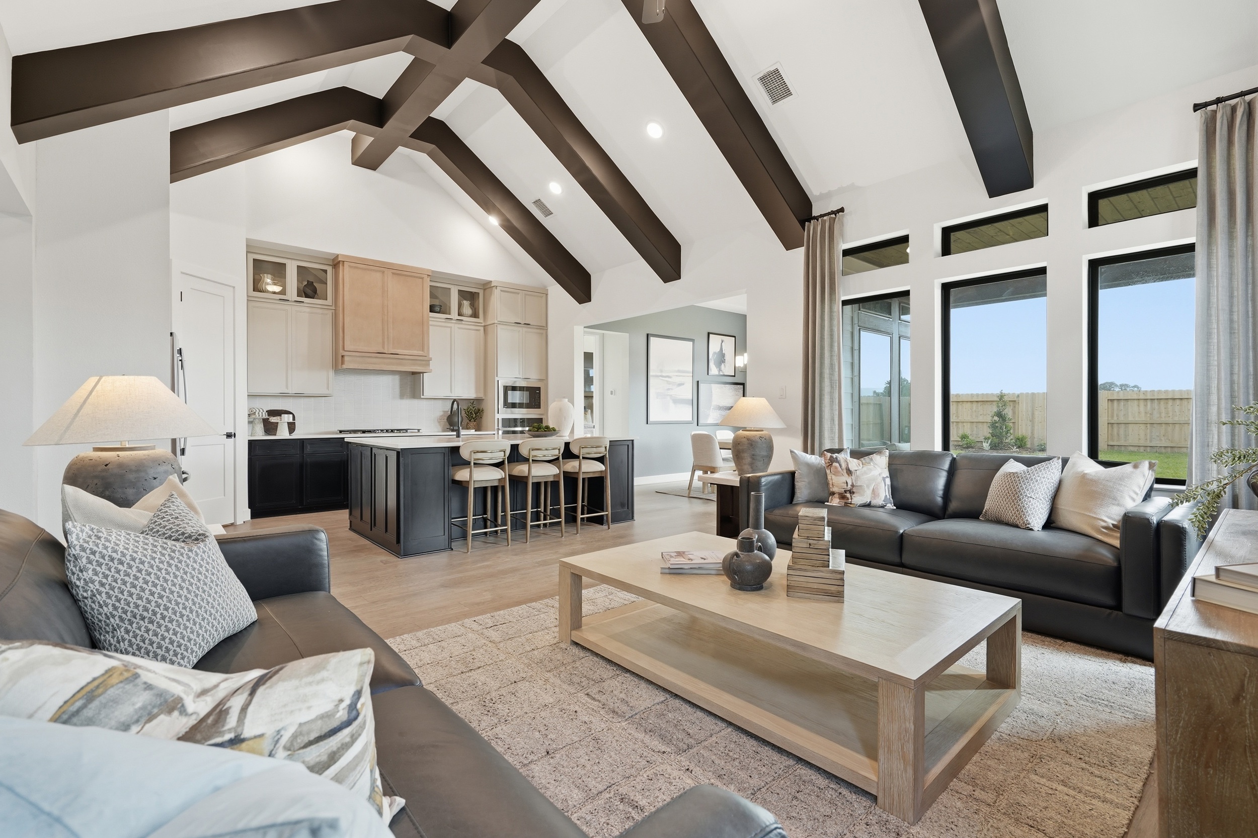 Spacious modern living room with vaulted ceilings, dark exposed beams, large windows, and an open kitchen—an inviting feature of New Homes in Rosharon, TX.