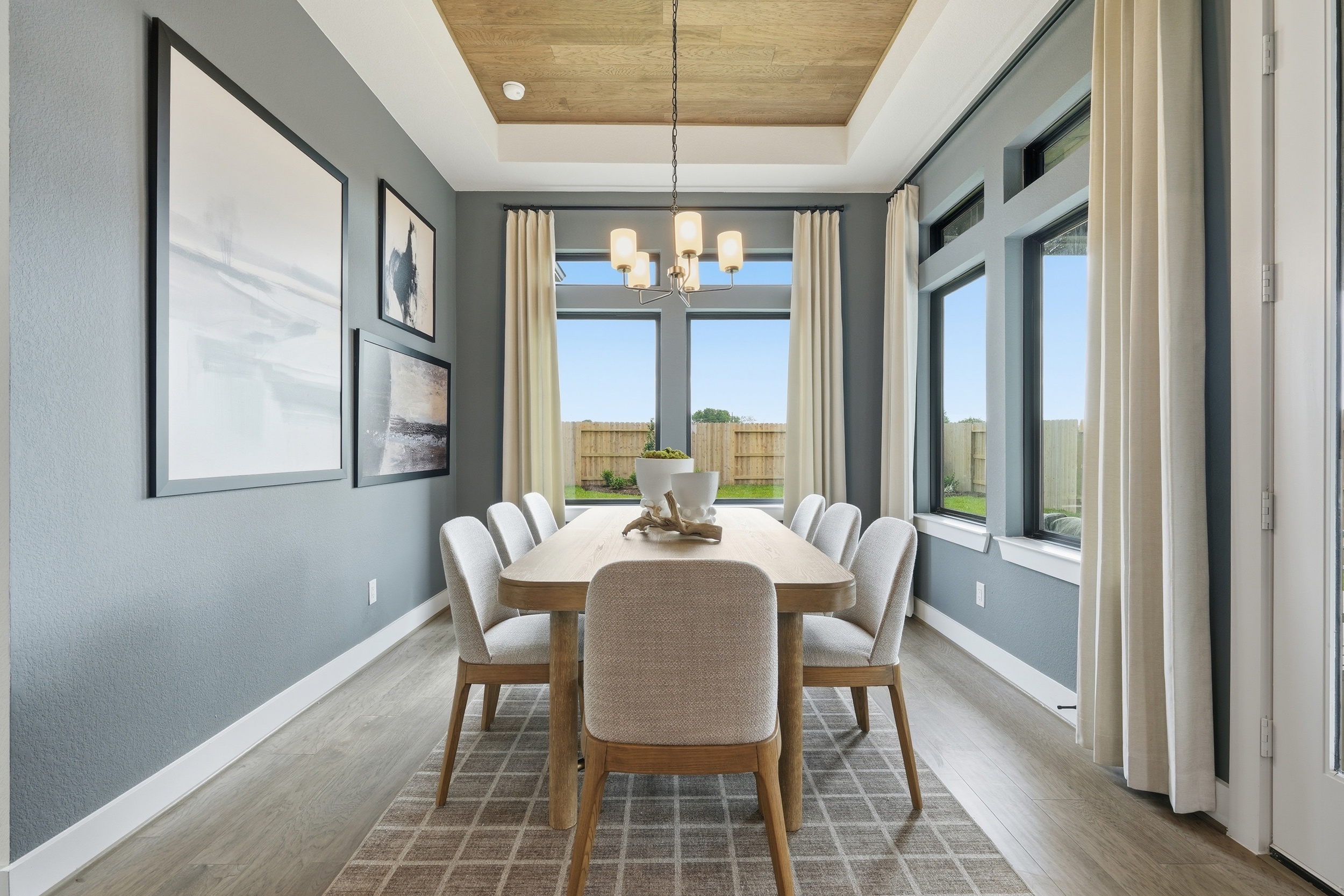 Modern dining room with a wooden table, six upholstered chairs, large windows with beige curtains, gray walls with artwork, a patterned rug, and a chandelier hanging from a wood-paneled ceiling—perfect for new homes in Rosharon, TX.
