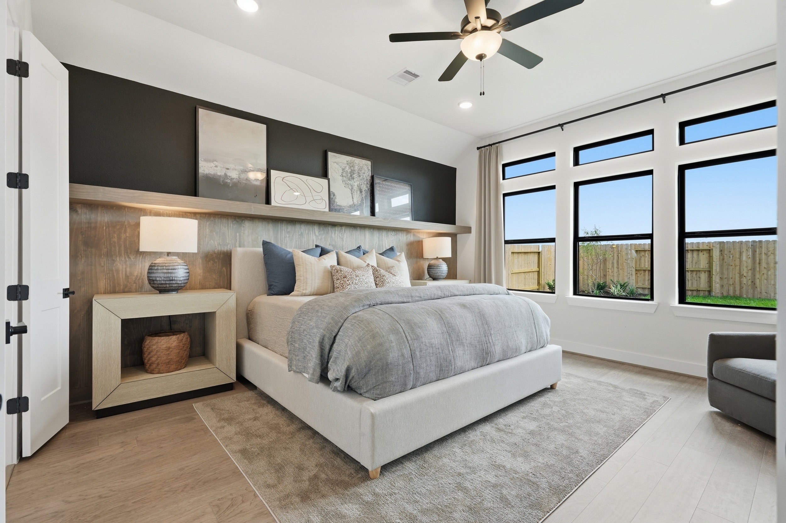 Modern bedroom in New Homes in Rosharon, TX, featuring a large bed with neutral bedding and blue accent pillows. A shelf above the bed displays framed art and lamps, while large windows fill the space with natural light. Ceiling fan included.