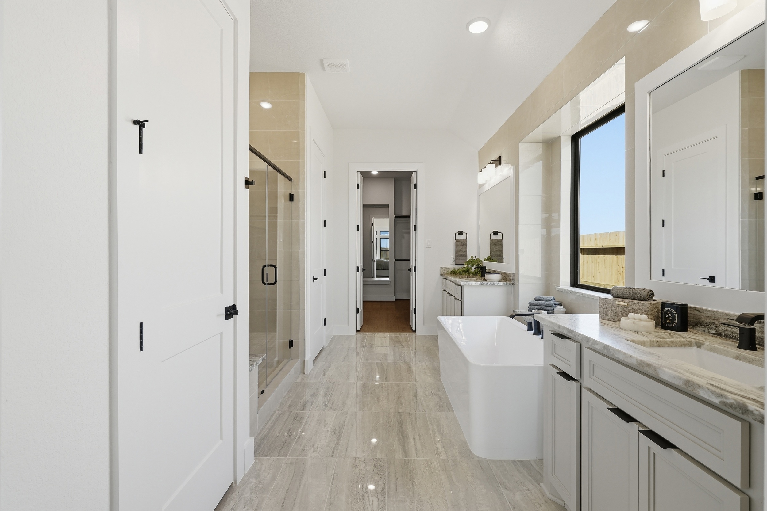 Modern bathroom with large tile floor, freestanding tub, glass shower, and dual-sink vanity. Enjoy natural light from a large window in this bright space with neutral tones—perfect for New Homes in Rosharon, TX.