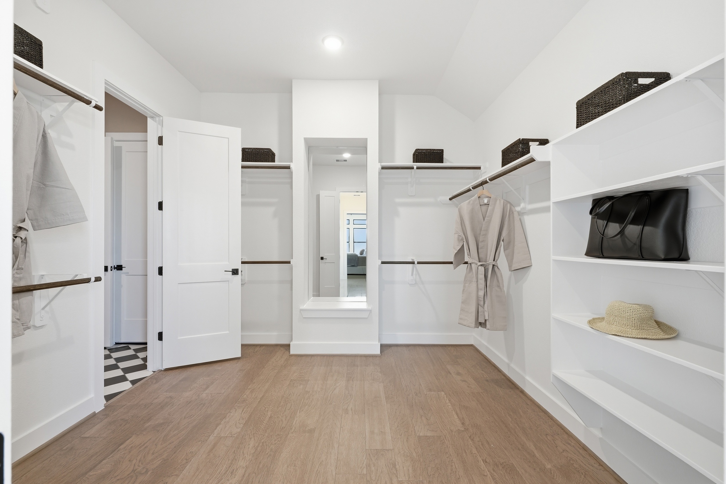 Spacious walk-in closet in one of the stunning New Homes in Rosharon, TX, featuring white shelves, hanging rods, storage baskets, a mirror, and a built-in bench. Light wood floors offer a warm touch to this elegant space.