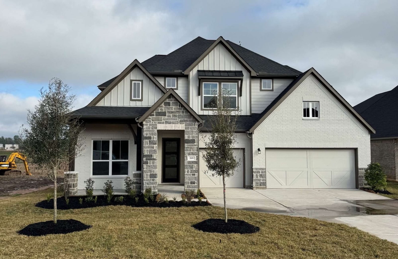 Two-story suburban house with a gray and white exterior, stone and brick detailing, a front porch, two-car garage, and a neatly landscaped yard with small trees and shrubs. A construction vehicle is visible in the background.