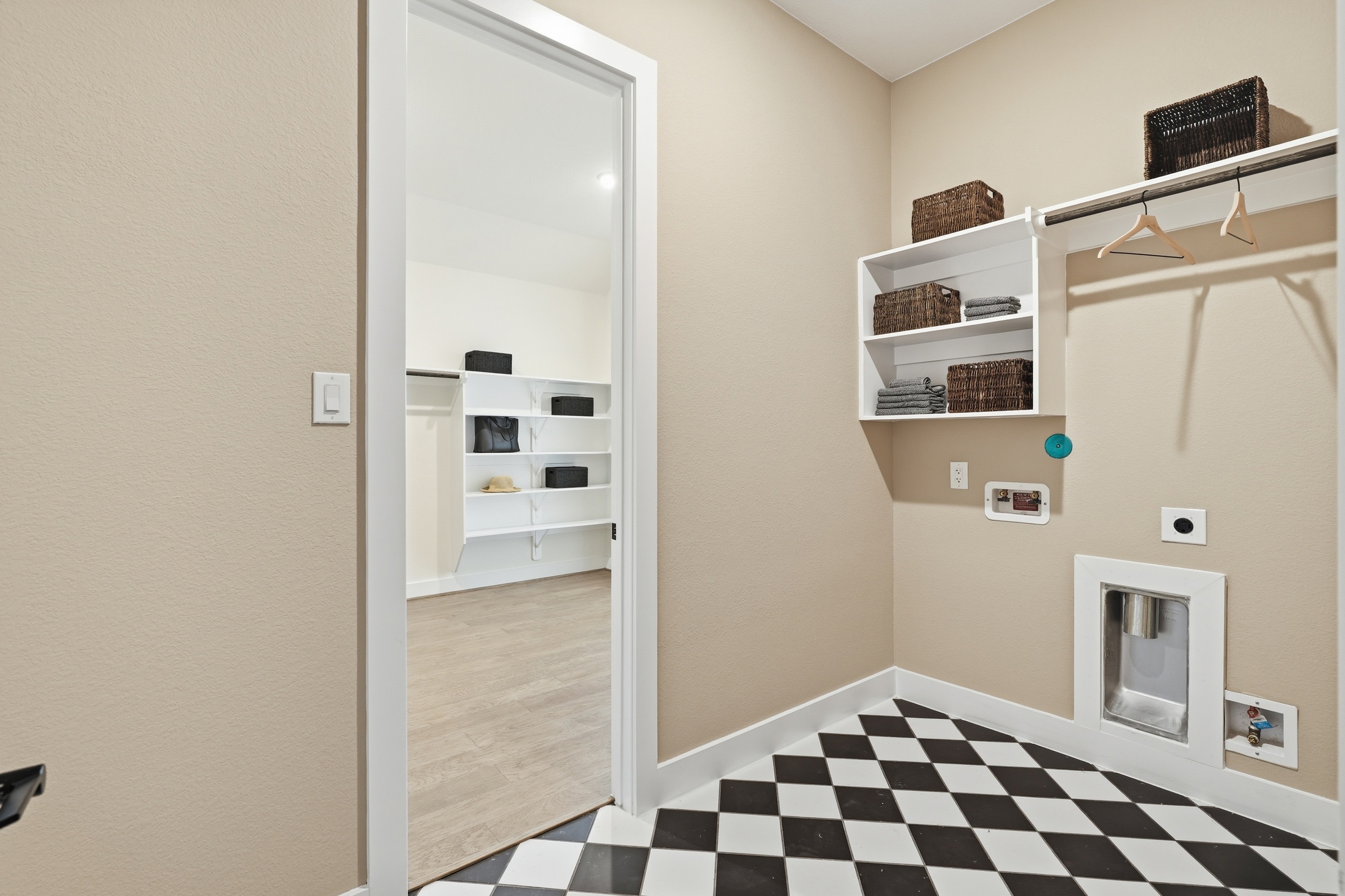 Laundry room in New Homes in Rosharon, TX features a black-and-white checkered floor, beige walls, white shelves with baskets and folded towels, wall hookups for washer and dryer, and a doorway to an adjacent room with extra shelving.
