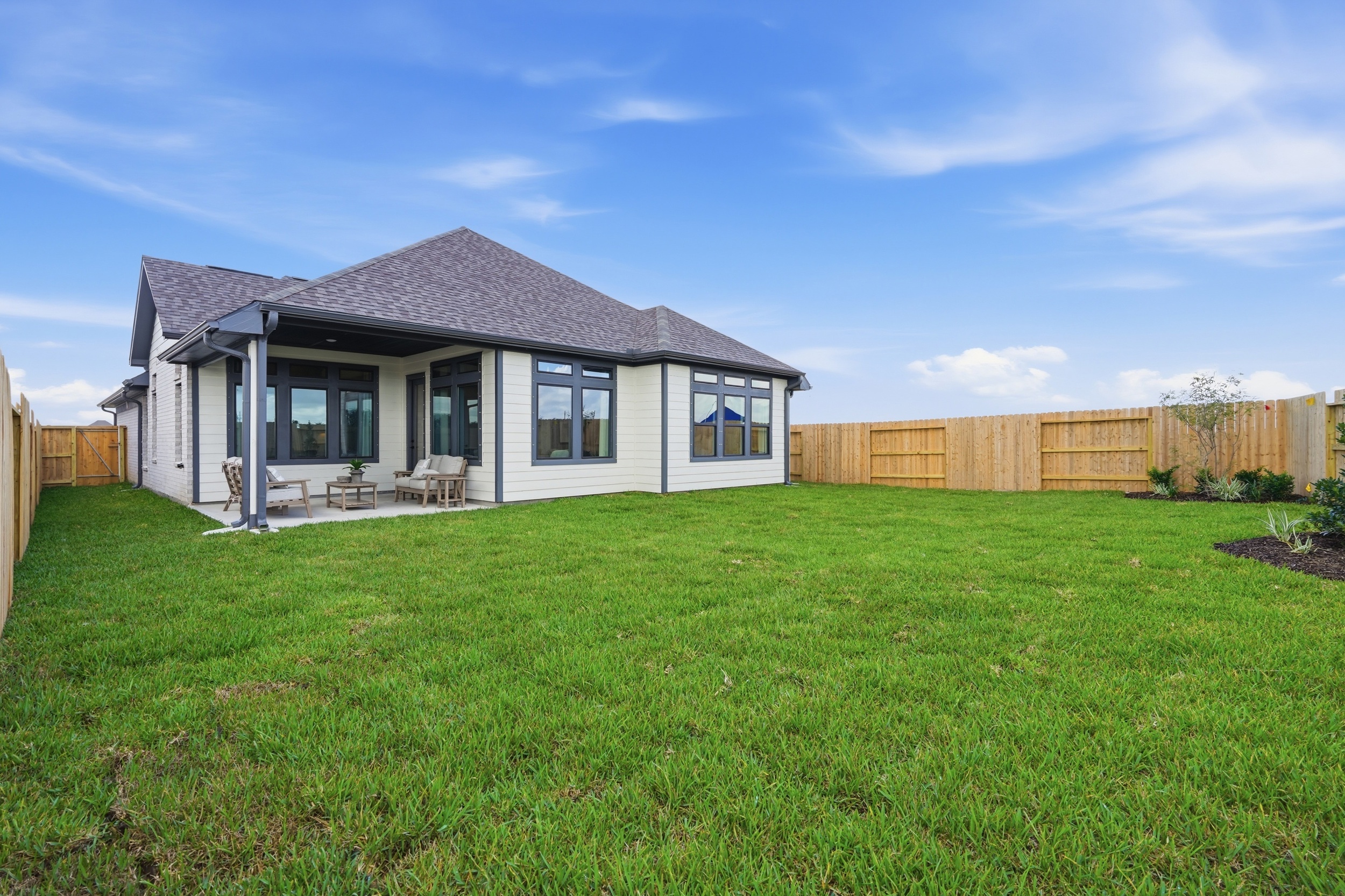 A modern single-story house with large windows sits in a spacious backyard in Rosharon, TX, featuring green grass, a covered patio with outdoor seating, and a wooden privacy fence under a blue sky—perfect for those seeking new homes in Rosharon.