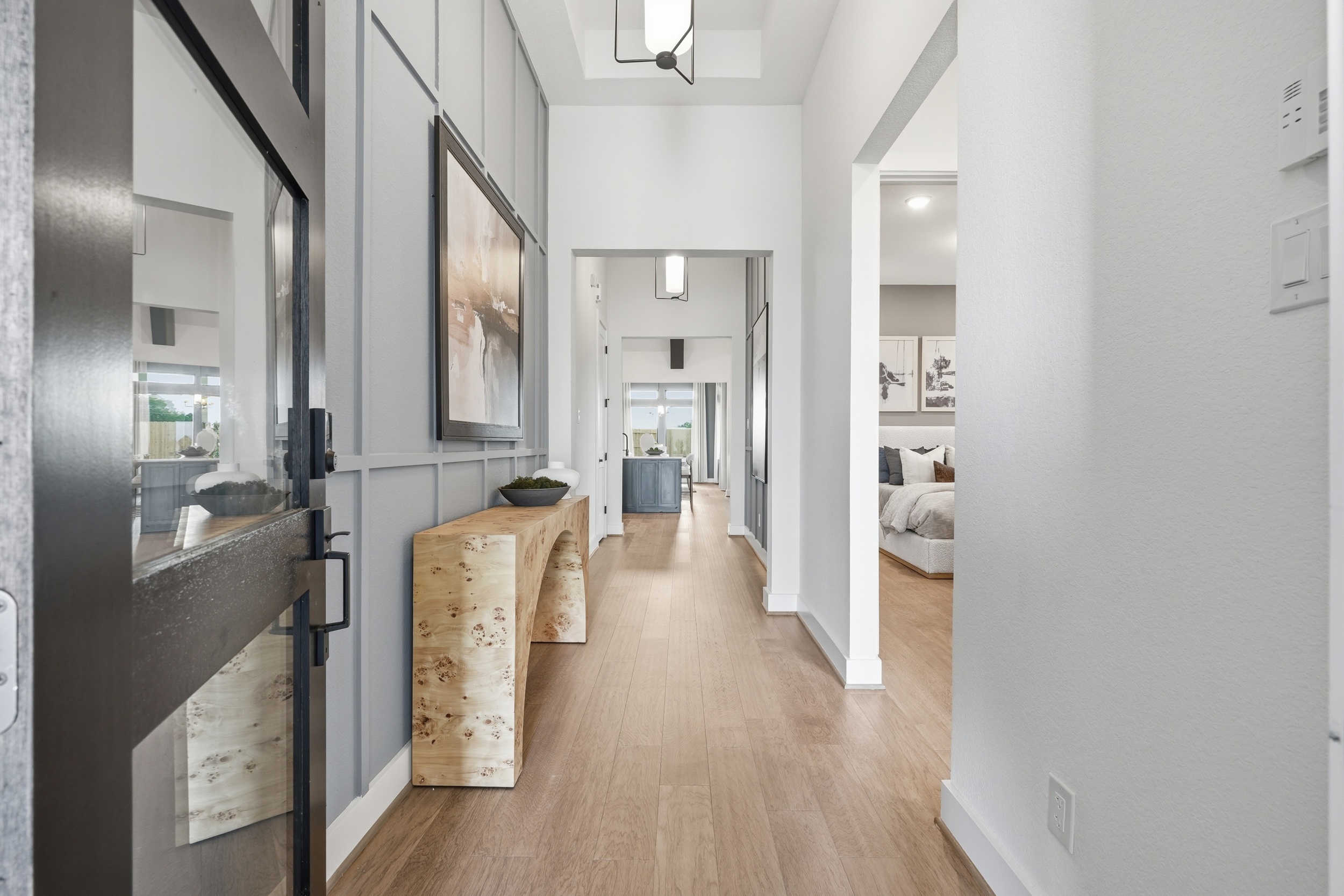 A modern hallway with light wood flooring, white walls, a wooden console table, framed artwork, and a glimpse into a bright living area and bedroom—perfectly showcasing the contemporary style of new homes in Rosharon, TX.