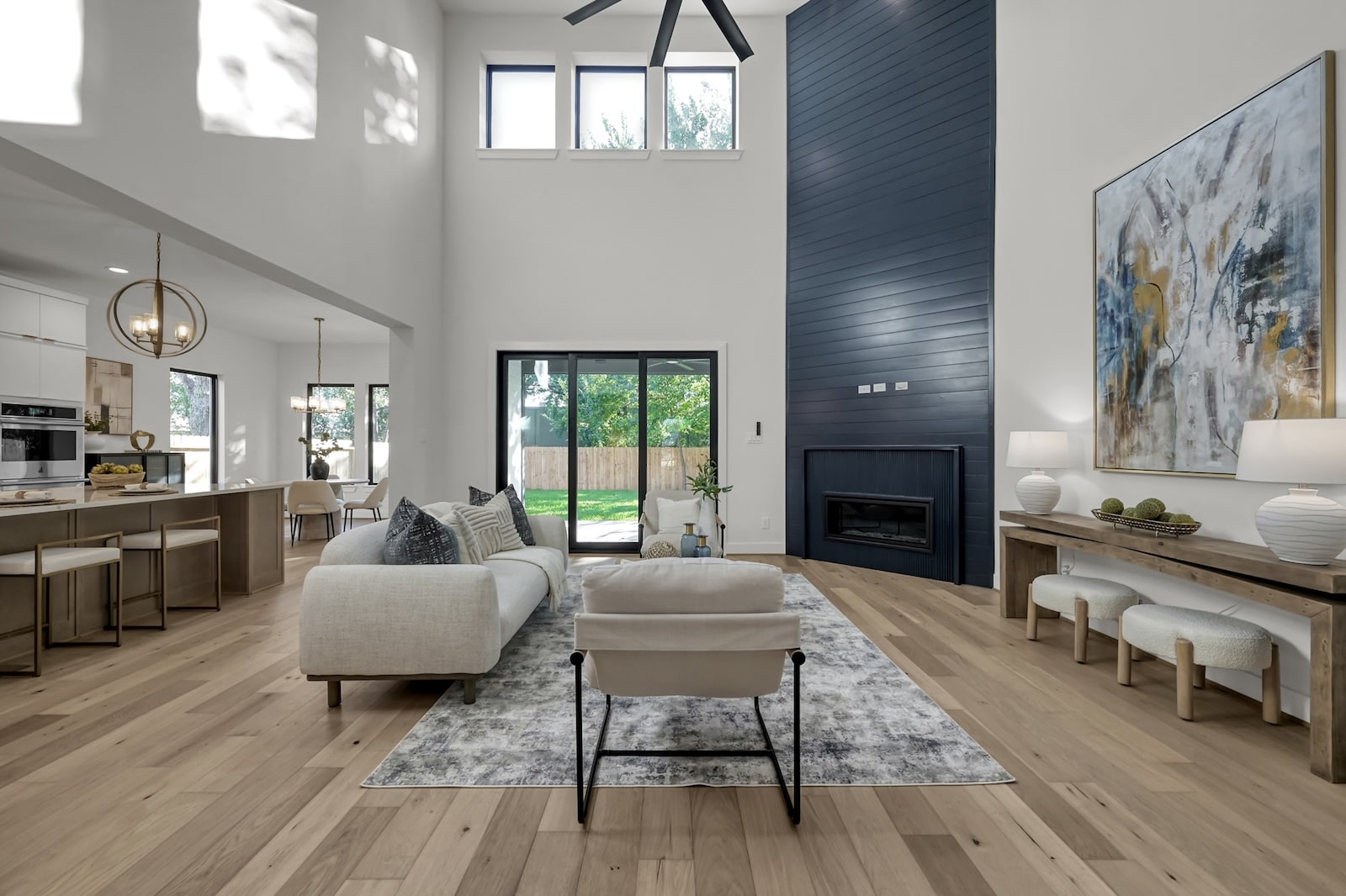 Modern living room with light wood floors, white walls, tall ceiling, large windows, and a dark accent fireplace. Neutral furniture, abstract art, and contemporary decor create a bright, open, and stylish space.