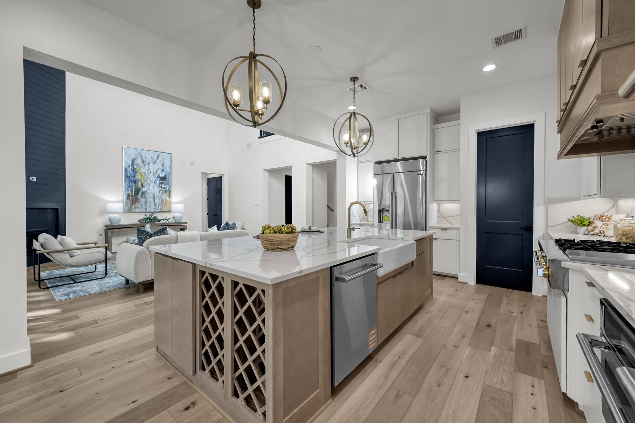 Modern kitchen with a large island featuring a built-in wine rack, pendant lights, stainless steel appliances, light wood floors, and an open view into a bright living room with a sofa and artwork.