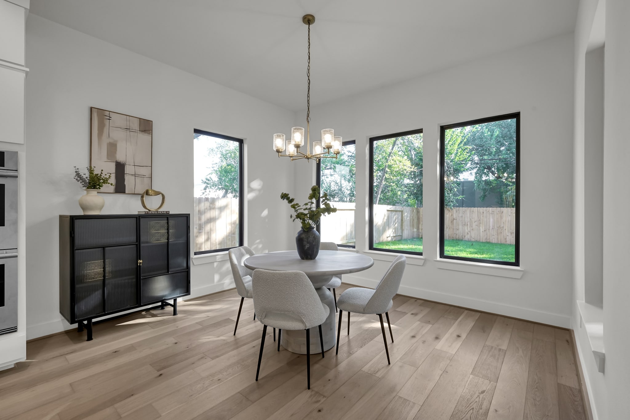 A modern dining area with a round white table, four cushioned chairs, a black side cabinet, minimalist decor, and three large windows showing a green backyard and wooden fence. Light wood flooring and a gold chandelier complete the space.