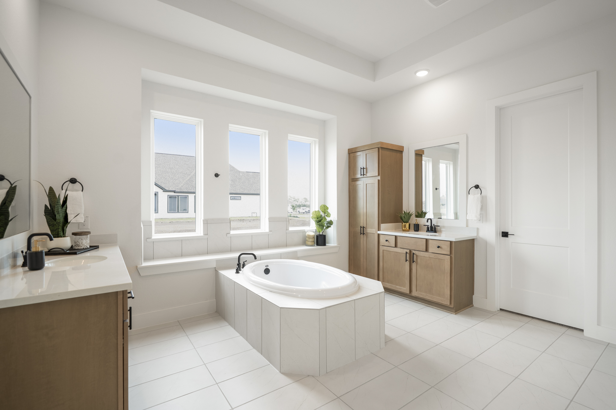 A bright, modern bathroom with a central white bathtub, large windows, wooden cabinets, two sinks, and potted plants on counters. White tile floors and walls create an airy, spacious feel.