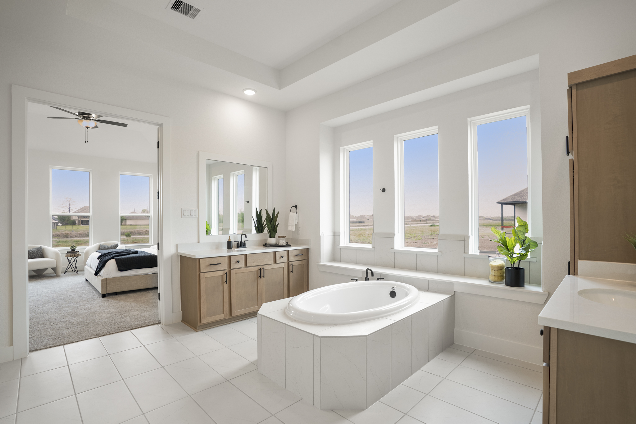 Spacious modern bathroom with a white bathtub set in the center, large windows, light wood cabinets, and green plants, leading into a bright bedroom with a large bed and ceiling fan.