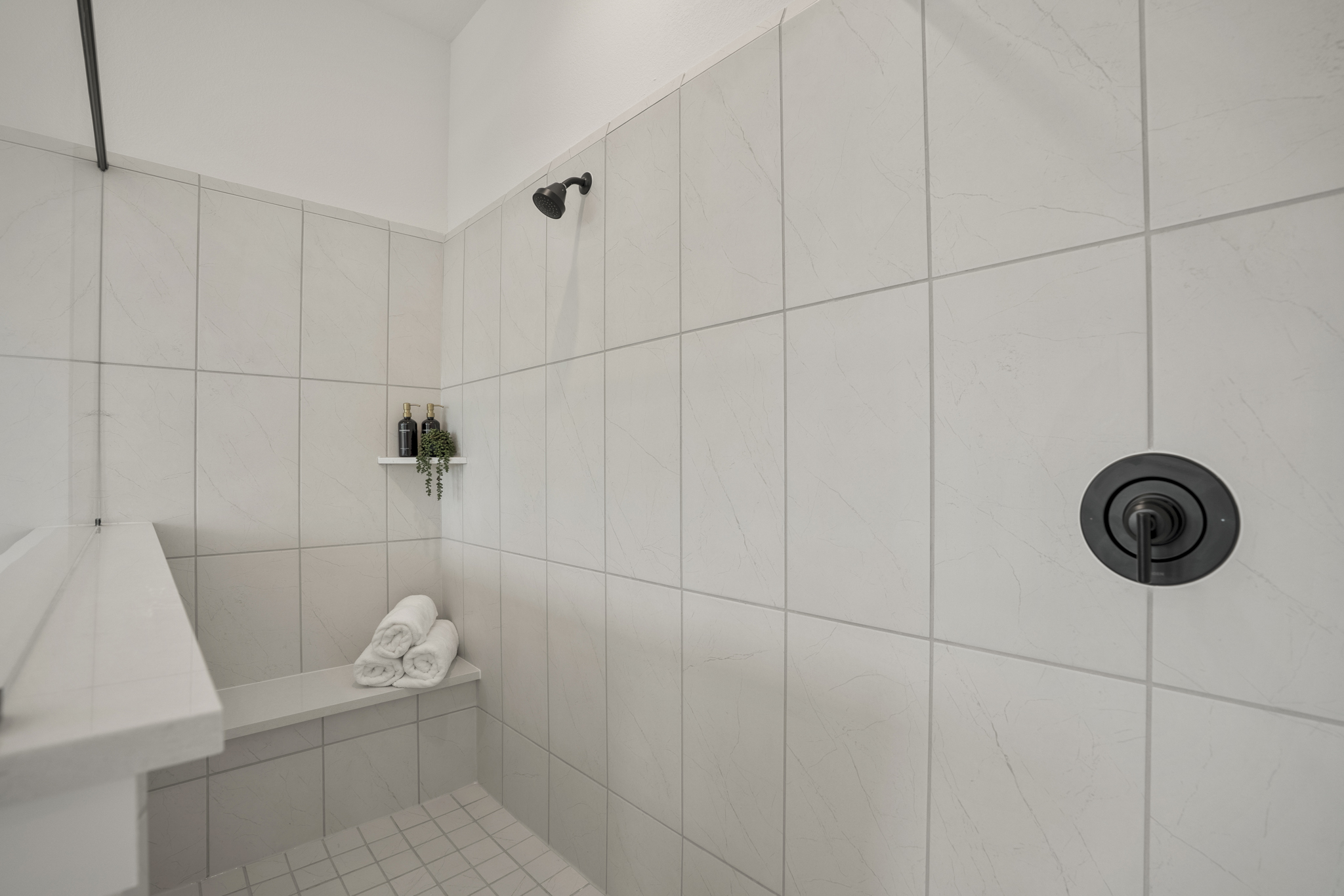 A modern shower with light-colored tile walls, a built-in bench holding rolled white towels, a small corner shelf with bottles and a plant, and matte black fixtures.
