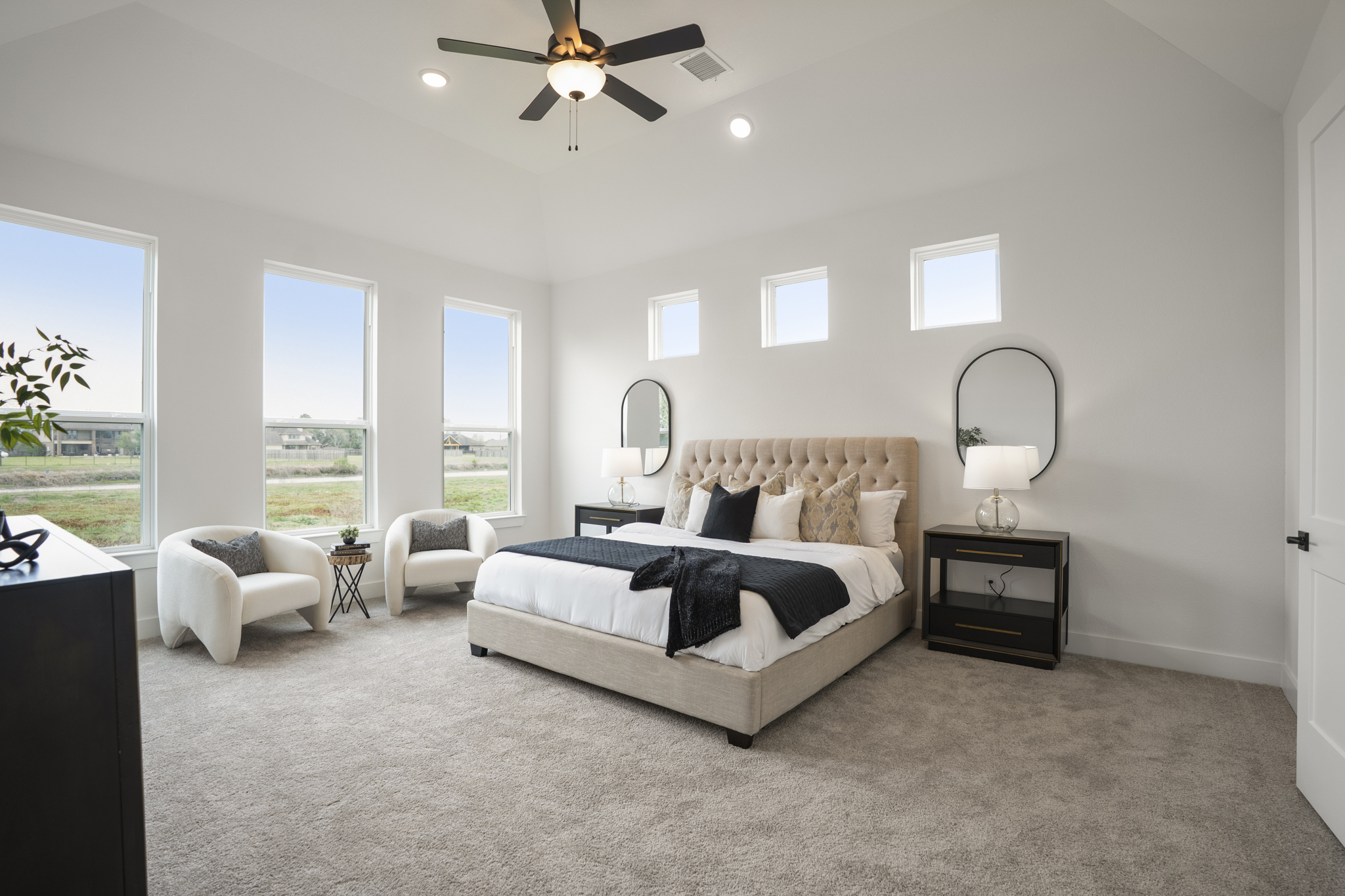 Modern bedroom with large windows, a beige tufted bed, white armchairs, black nightstands with lamps and round mirrors, light carpet, and a ceiling fan. The room is bright and decorated in neutral tones.