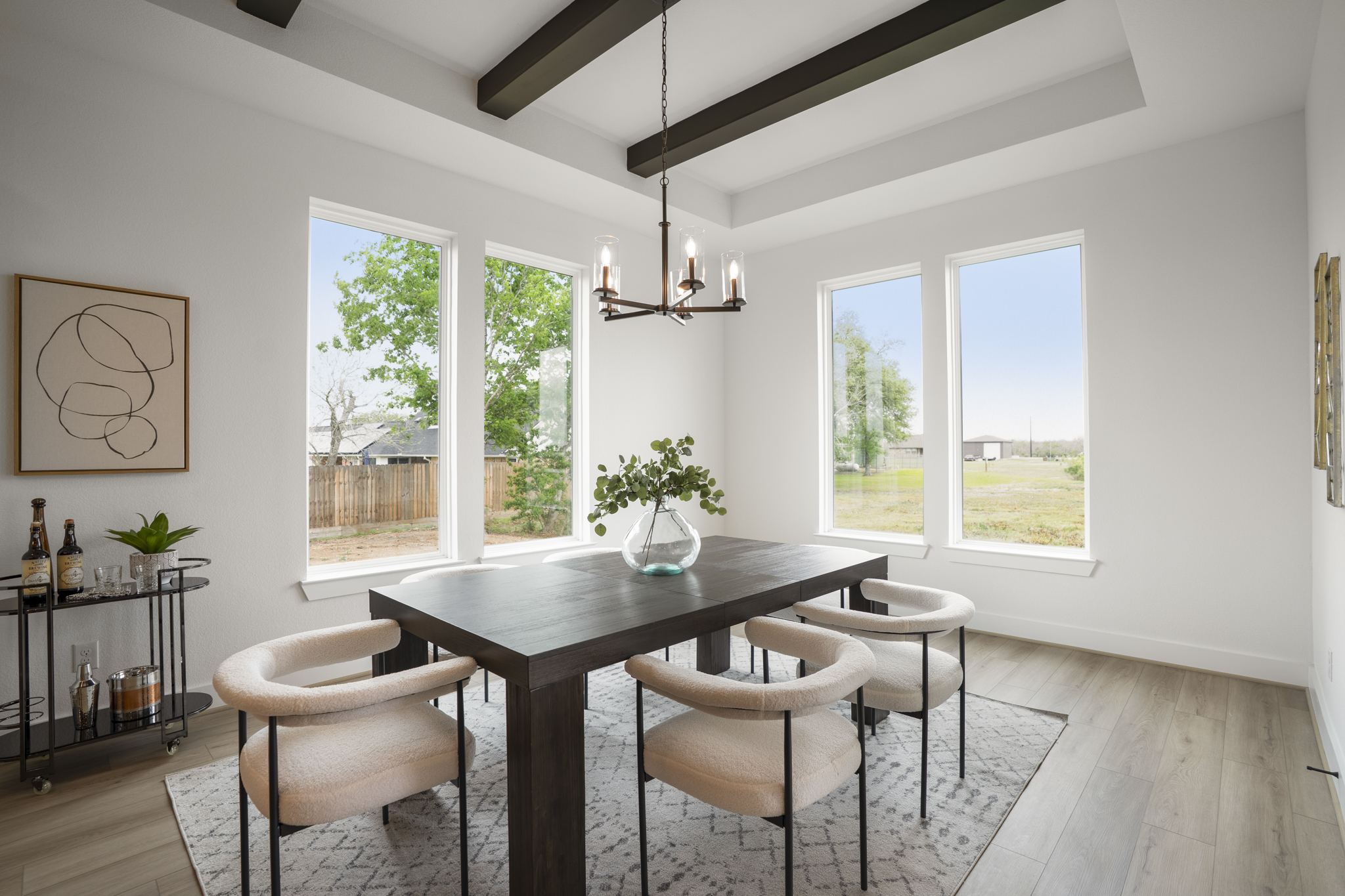 A modern dining room with large windows, a dark wood table, six light upholstered chairs, a geometric rug, a glass vase with greenery, a bar cart, and a contemporary chandelier under exposed ceiling beams.