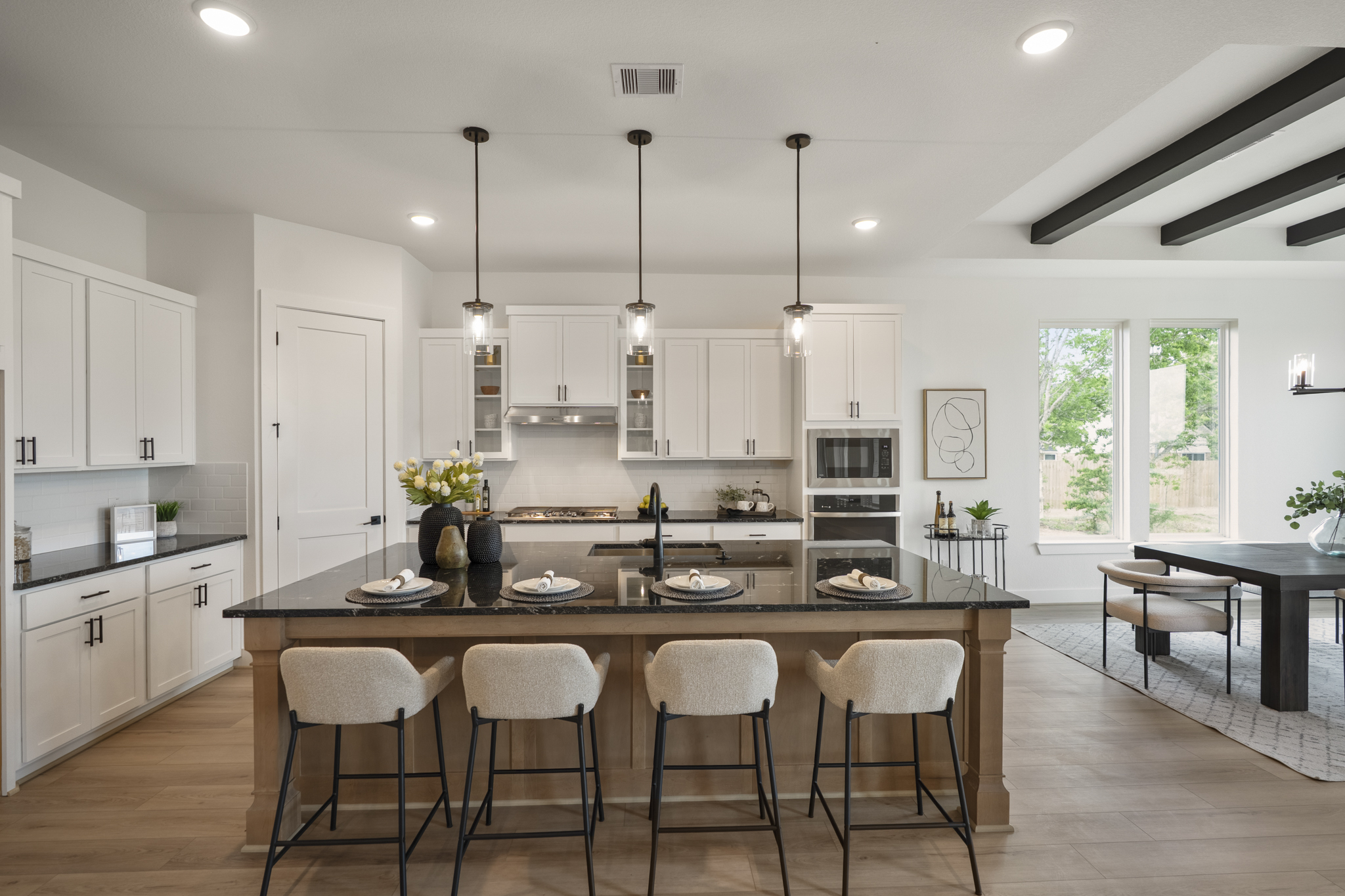 Modern kitchen with white cabinets, black granite island with four beige barstools, hanging pendant lights, and adjacent dining area with large windows letting in natural light.