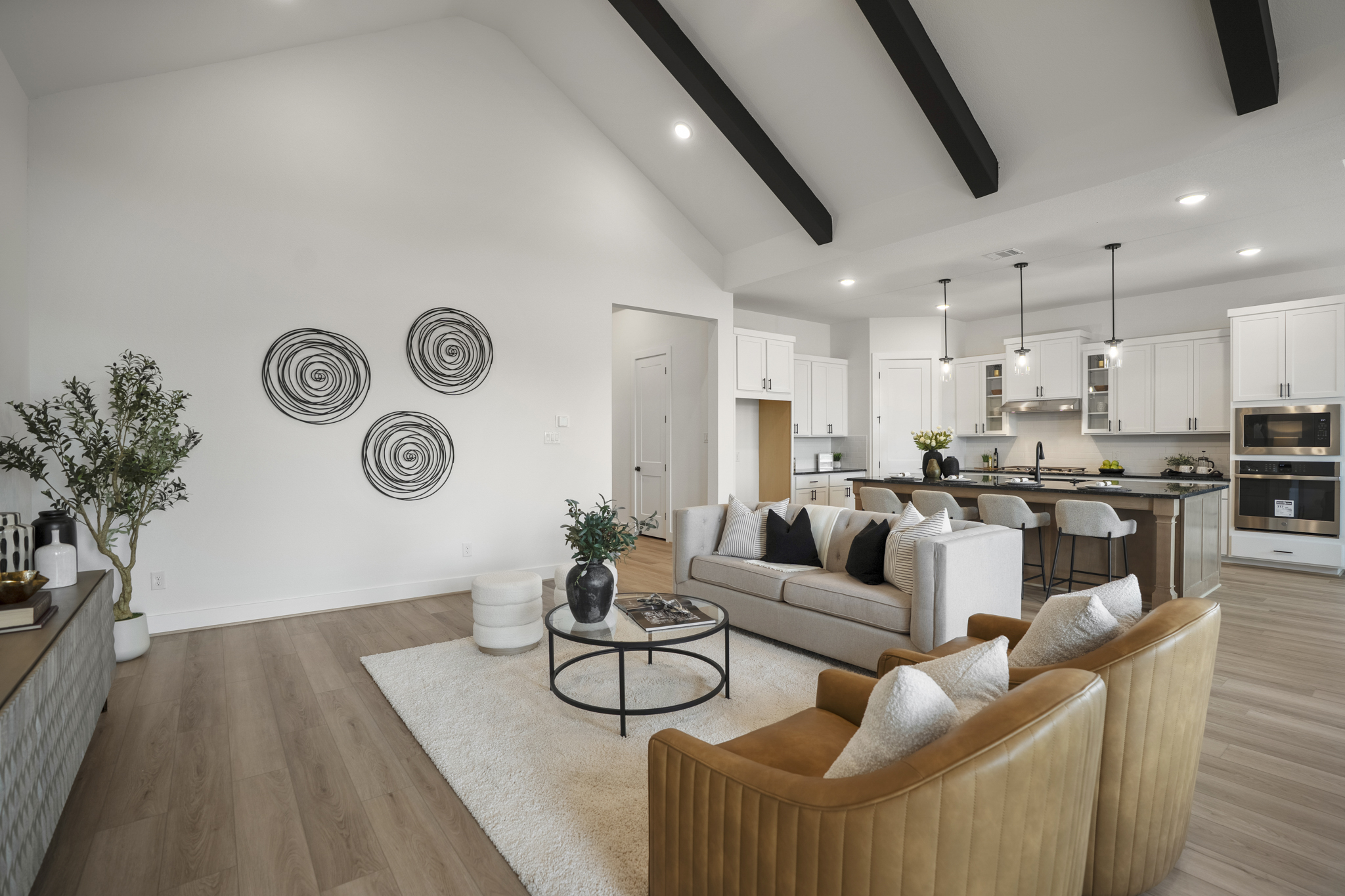 Modern open-concept living room and kitchen with vaulted ceiling, wood beams, neutral decor, tan chairs, a light sofa, round coffee table, wall art, and large kitchen island with pendant lighting.