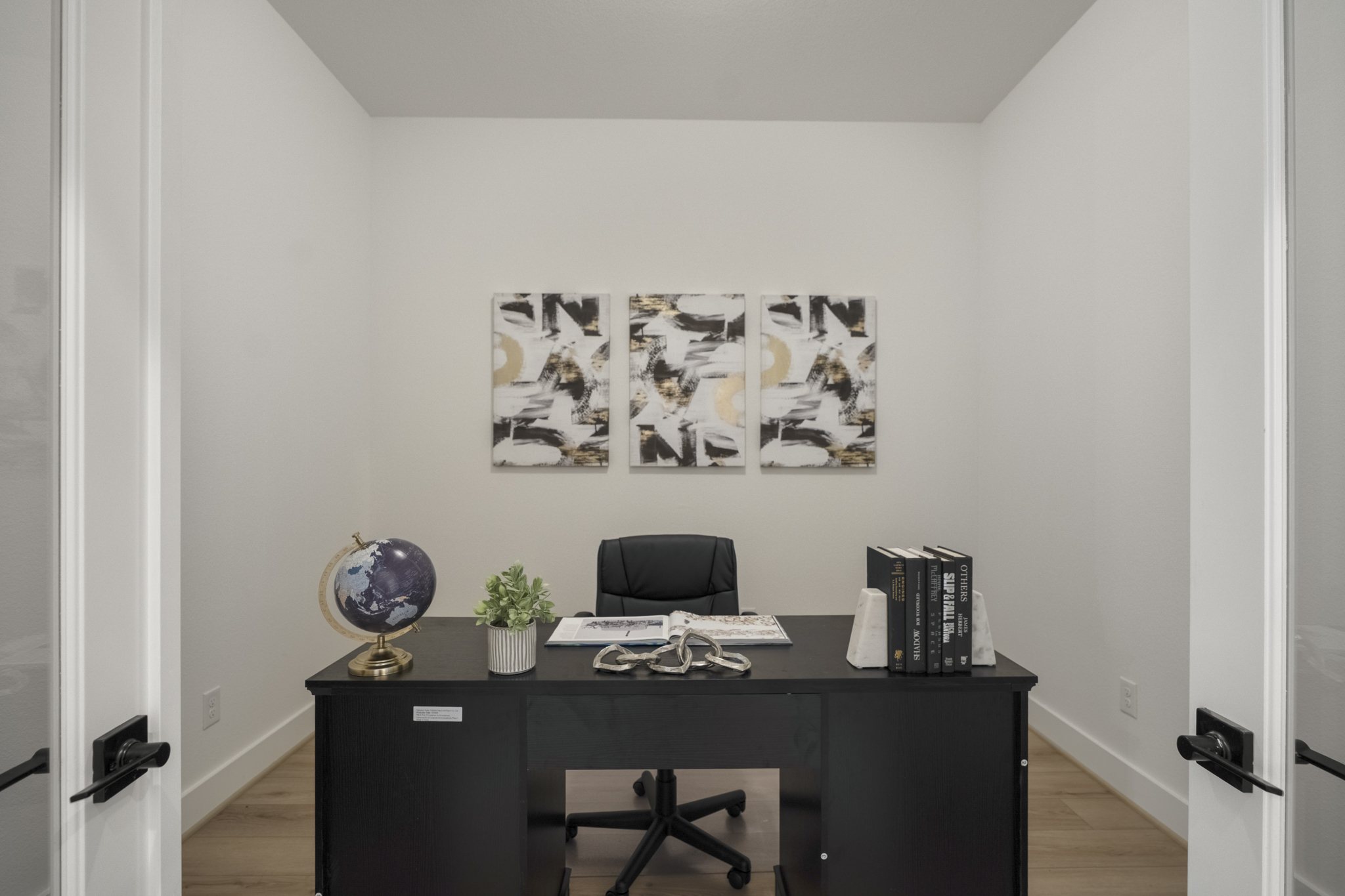 A modern, minimalist home office with a black desk, black chair, globe, potted plant, books, and decorative items. Three abstract paintings hang on the white wall behind the desk. Light wood floor and double doors are visible.