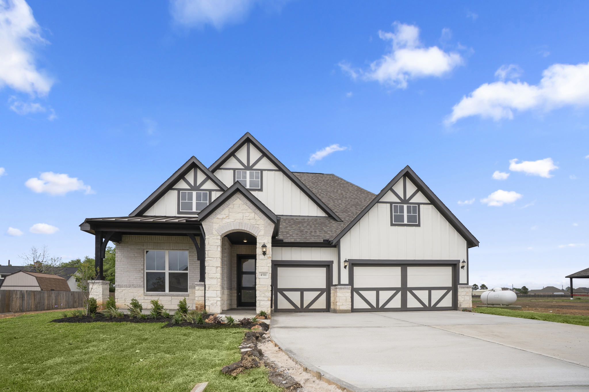 Modern suburban house with a white exterior, gray trim, and a three-car garage, set against a bright blue sky with scattered clouds. Fresh landscaping and a wide driveway are visible in front.