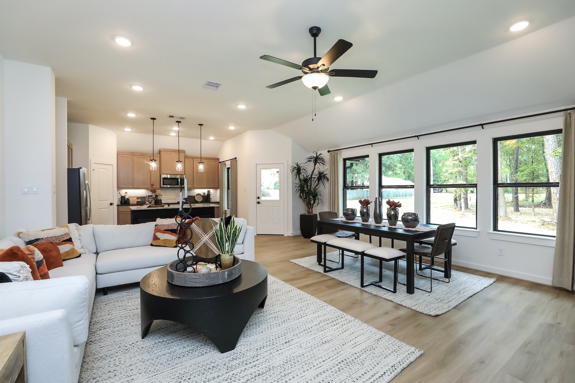 Modern open-concept living and dining area with large windows, a white sectional sofa, round black coffee table, dining table for six, pendant lights, ceiling fan, and light wood floors. Kitchen visible in the background.