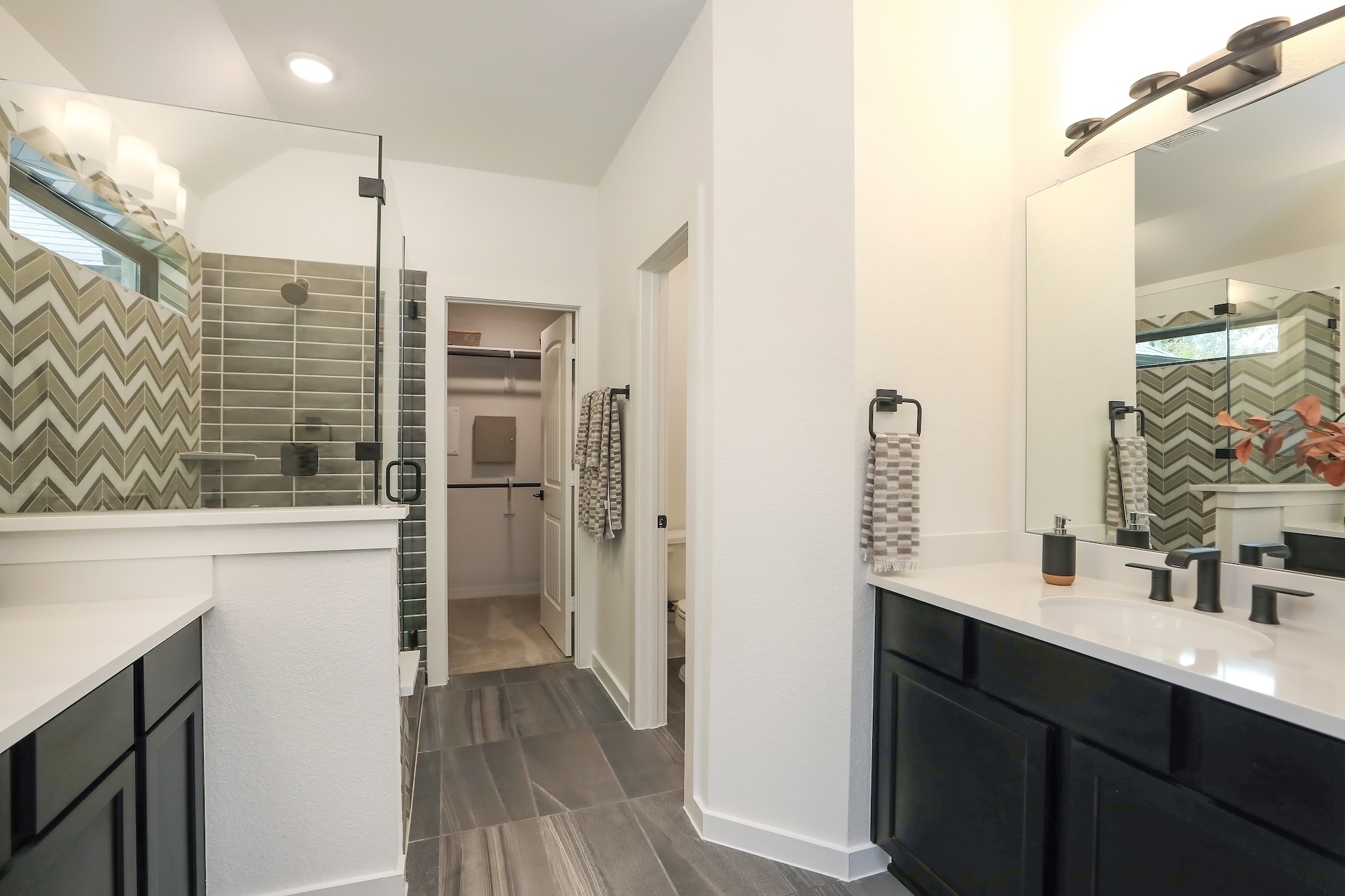 Modern bathroom with dark cabinets, white countertops, a large mirror, glass shower with chevron-patterned tiles, gray floor tiles, and neatly hung towels. Visible closet through open door in the back.