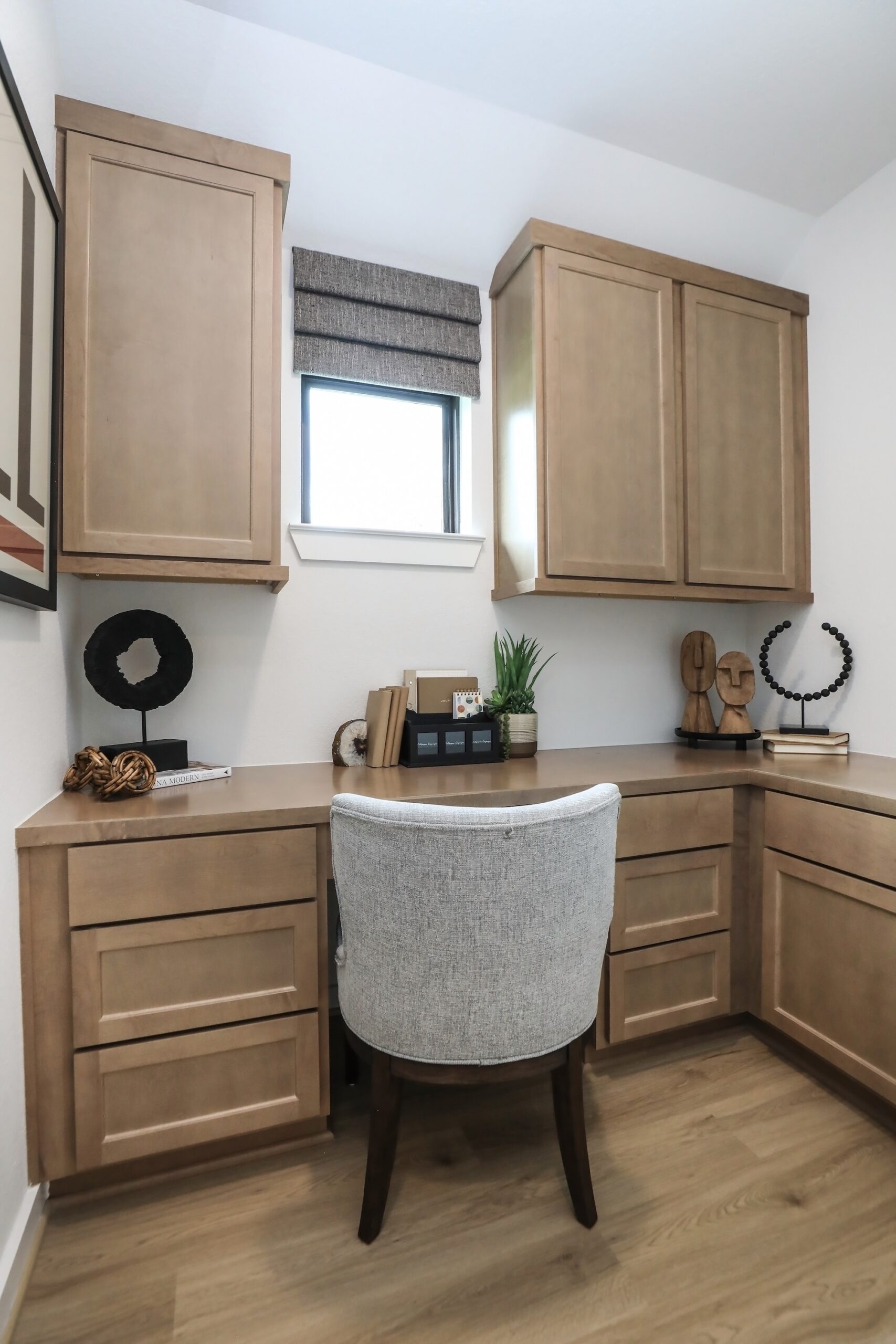 A modern home office with light wood cabinets, a gray cushioned chair, a small window with a brown shade, and decorative accents on the L-shaped desk, including books, plants, and abstract sculptures.