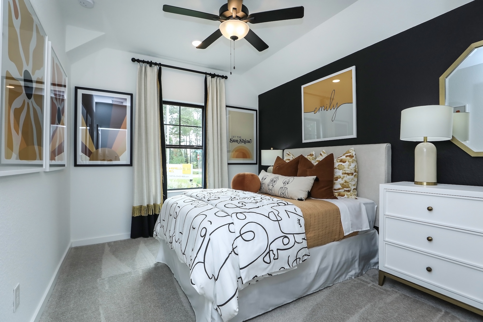 A modern bedroom with a black accent wall, framed art, a bed with white and brown bedding, decorative pillows, a white dresser with a lamp, and a window with black curtains letting in natural light.