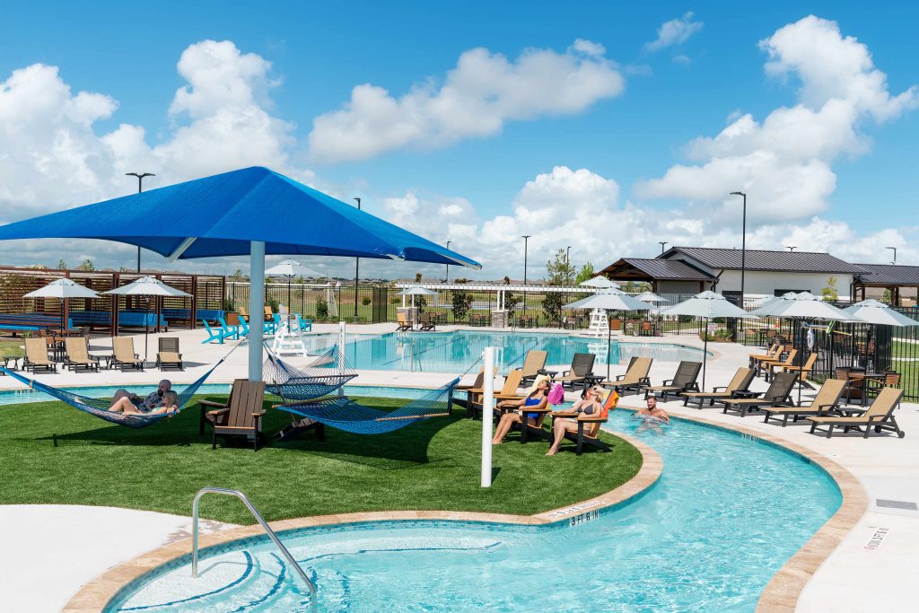 A bright outdoor pool area with lounge chairs, umbrellas, hammocks, and a shaded seating area. People are relaxing by the pool, in hammocks, and sitting by the water under a partly cloudy blue sky.