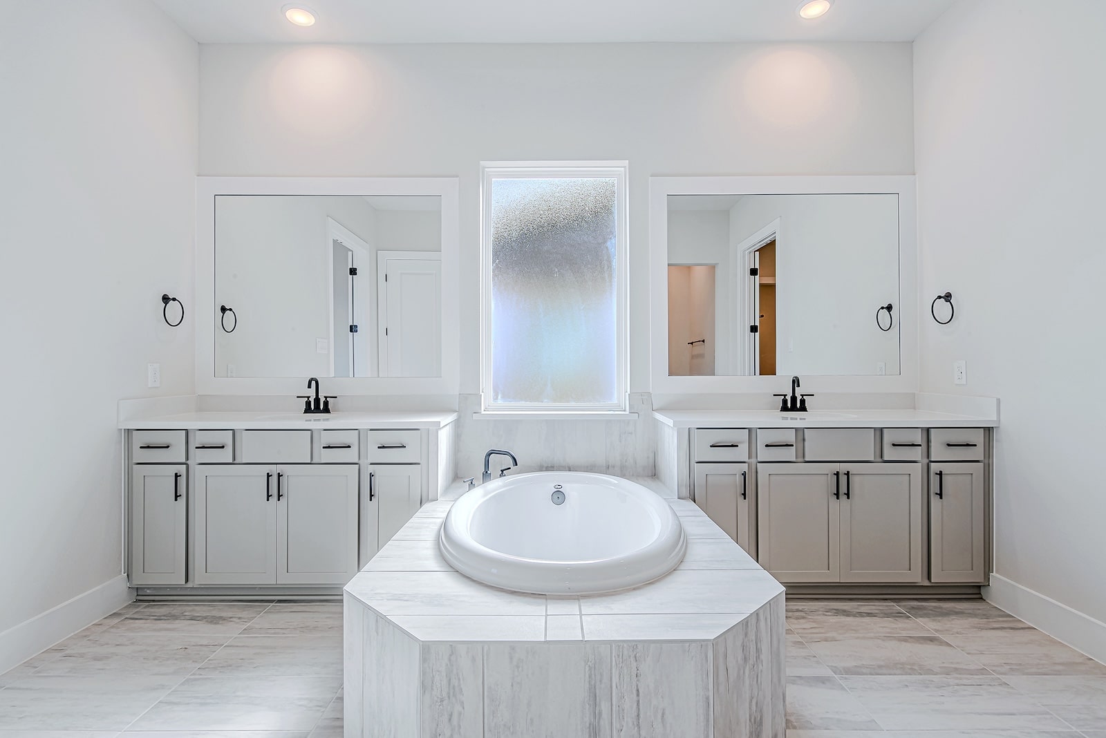 Modern bathroom with a central round bathtub, two large mirrors above matching vanities with sinks, and a frosted window between them. Light gray cabinetry and tile flooring create a clean, bright look.
