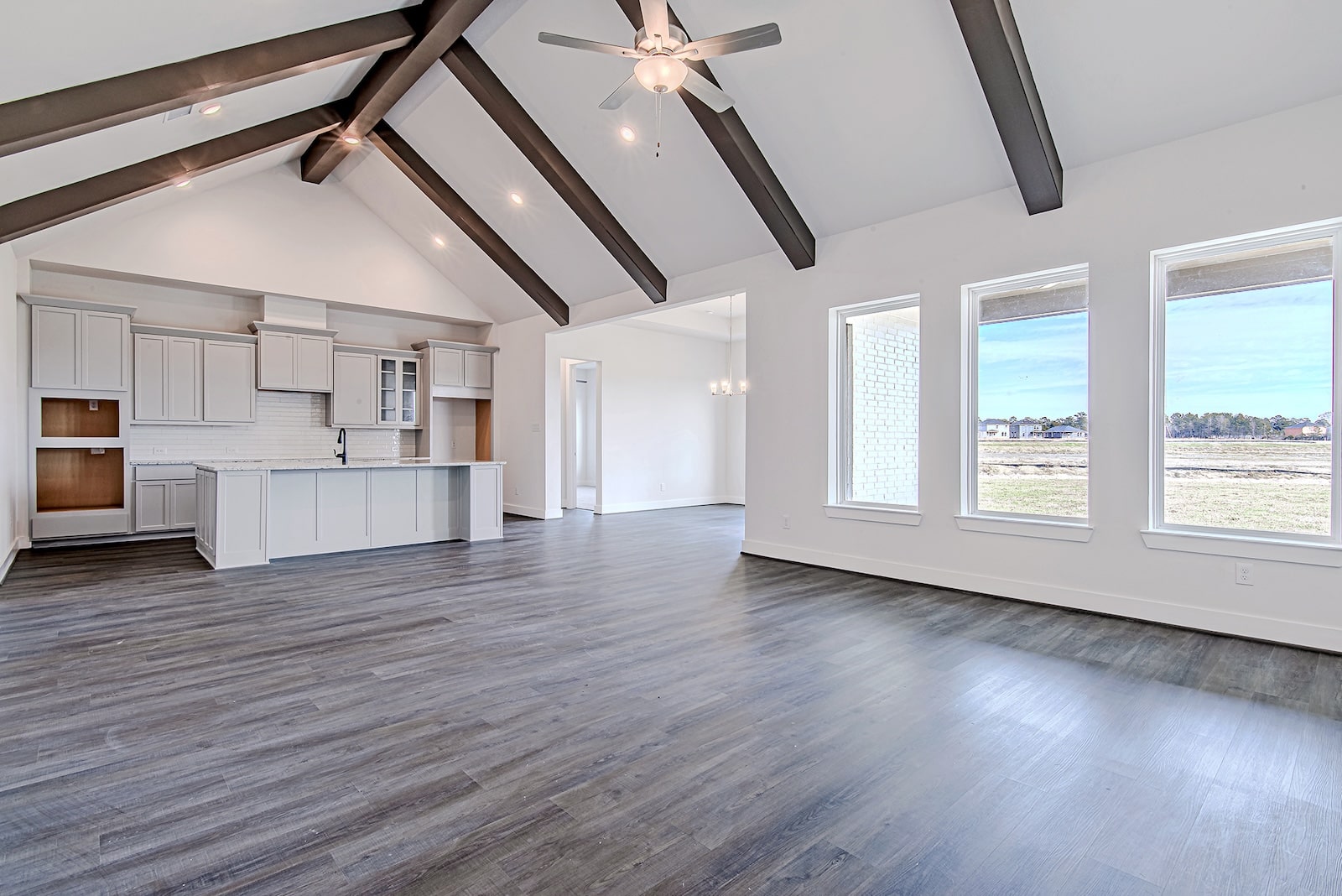 Spacious, modern open-concept living area with high vaulted ceiling, dark wooden beams, ceiling fan, large windows, and light wood floors. White kitchen with an island is visible in the background.