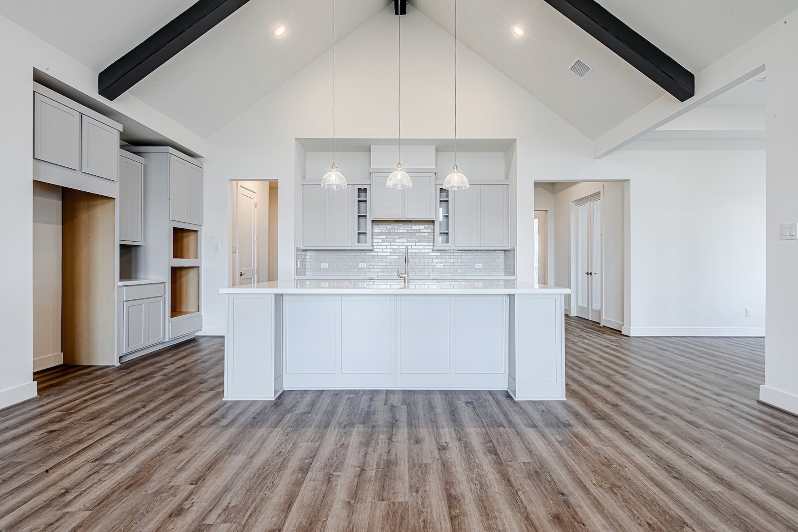 Modern, spacious kitchen with vaulted ceiling, white cabinetry, pendant lights over a large island, subway tile backsplash, and wood-style flooring. The room is bright with natural light and has an open layout.
