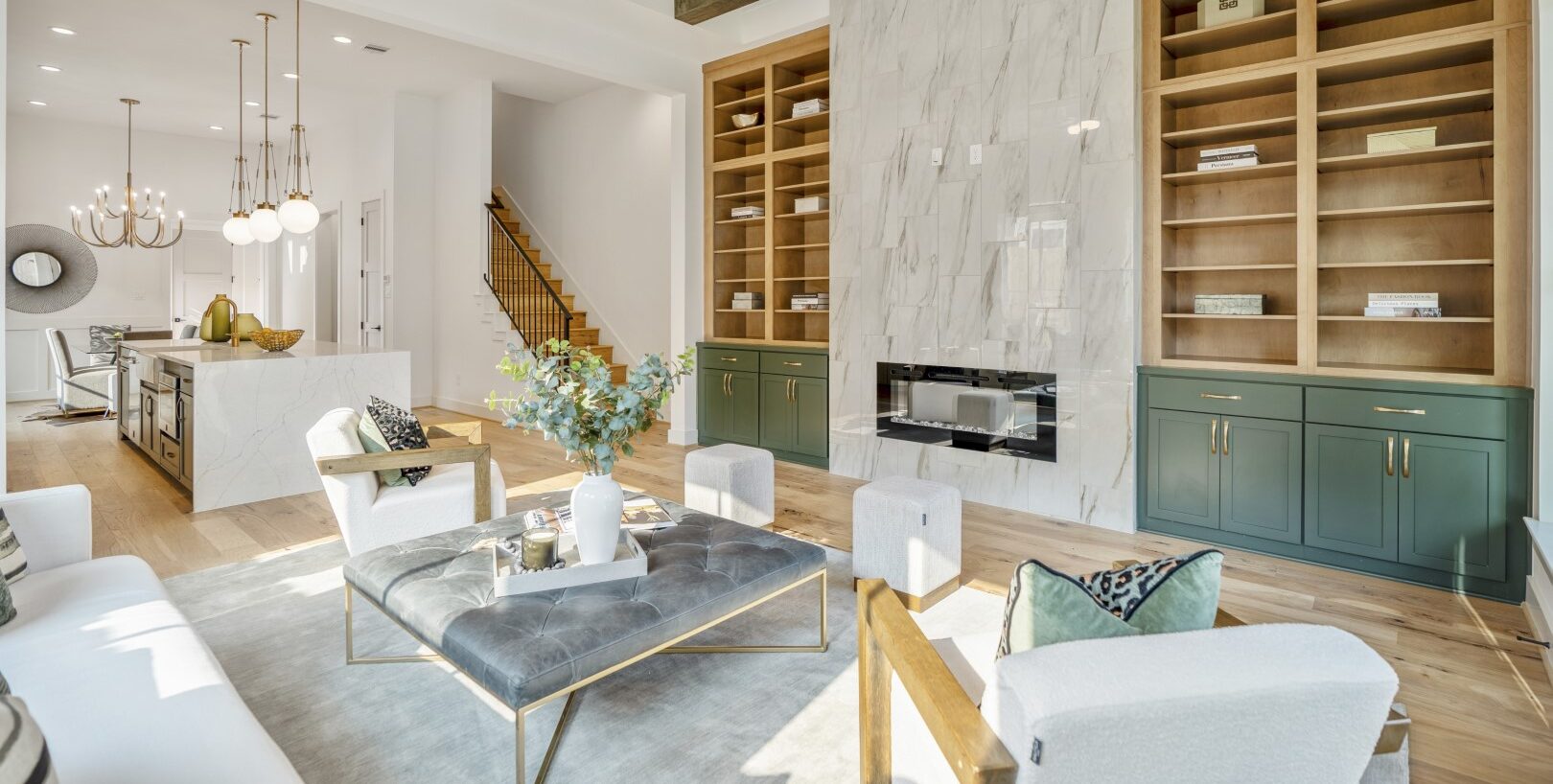 Modern open-concept living room with a marble fireplace, built-in wooden shelves, green cabinets, a tufted ottoman coffee table, white armchairs, and a kitchen with pendant lights in the background.