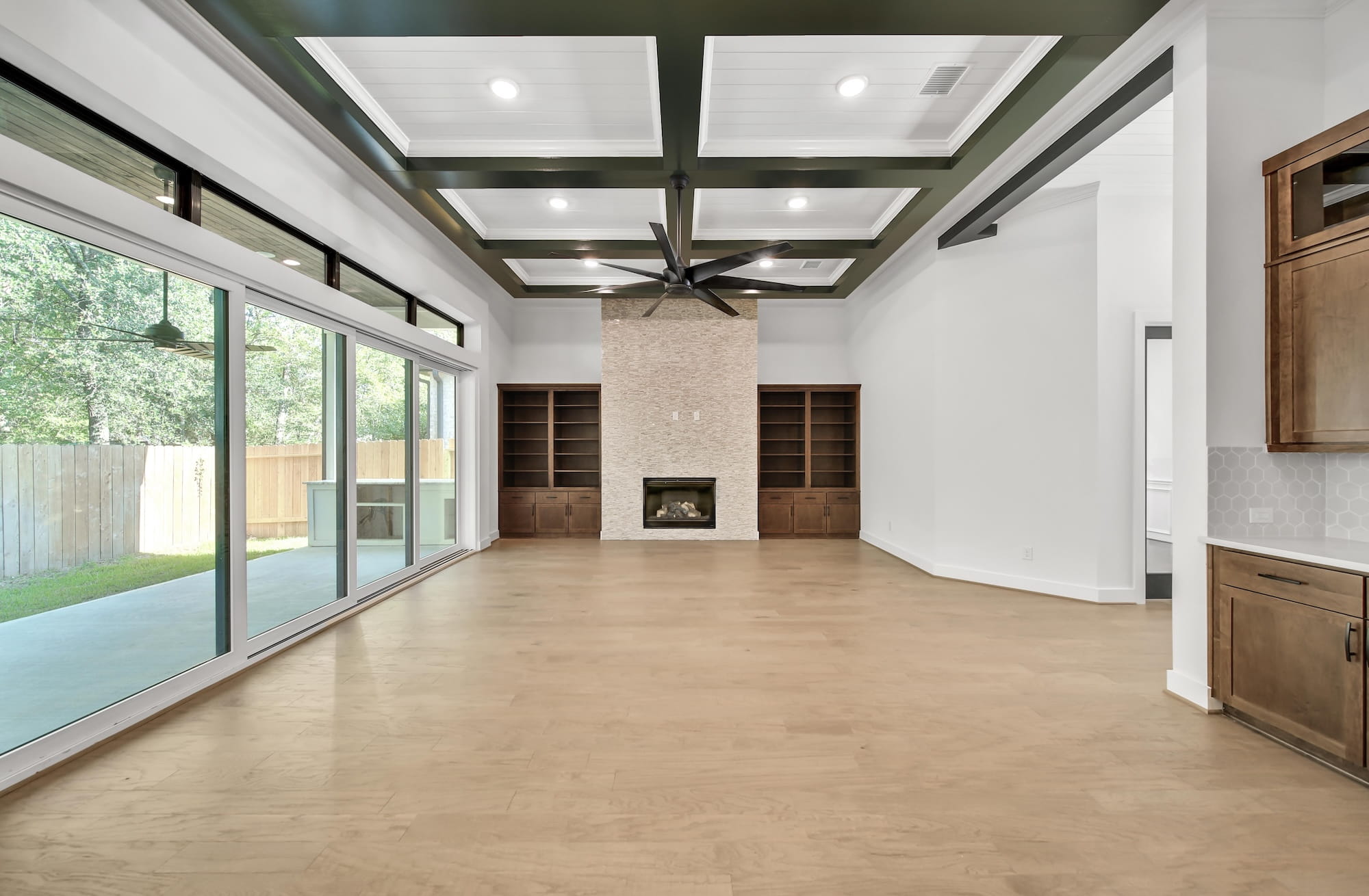 Spacious living room with large sliding glass doors, wood floors, built-in shelves, modern fireplace, coffered ceiling with recessed lighting, and view of a covered patio and fenced backyard.