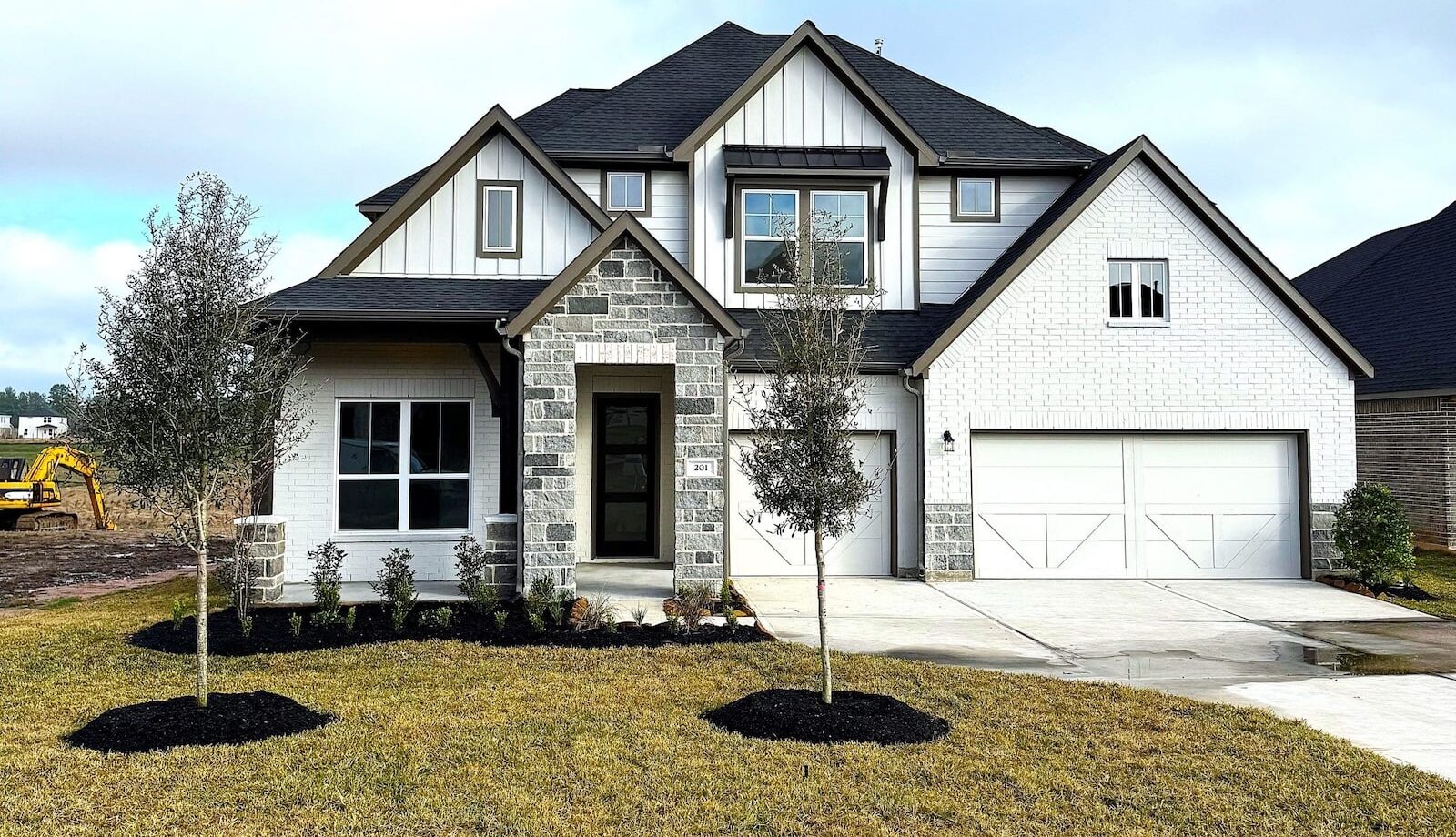 Two-story modern suburban house with white brick and gray stone accents, black roof, double garage, front porch, and three young trees planted in the front yard on a cloudy day.