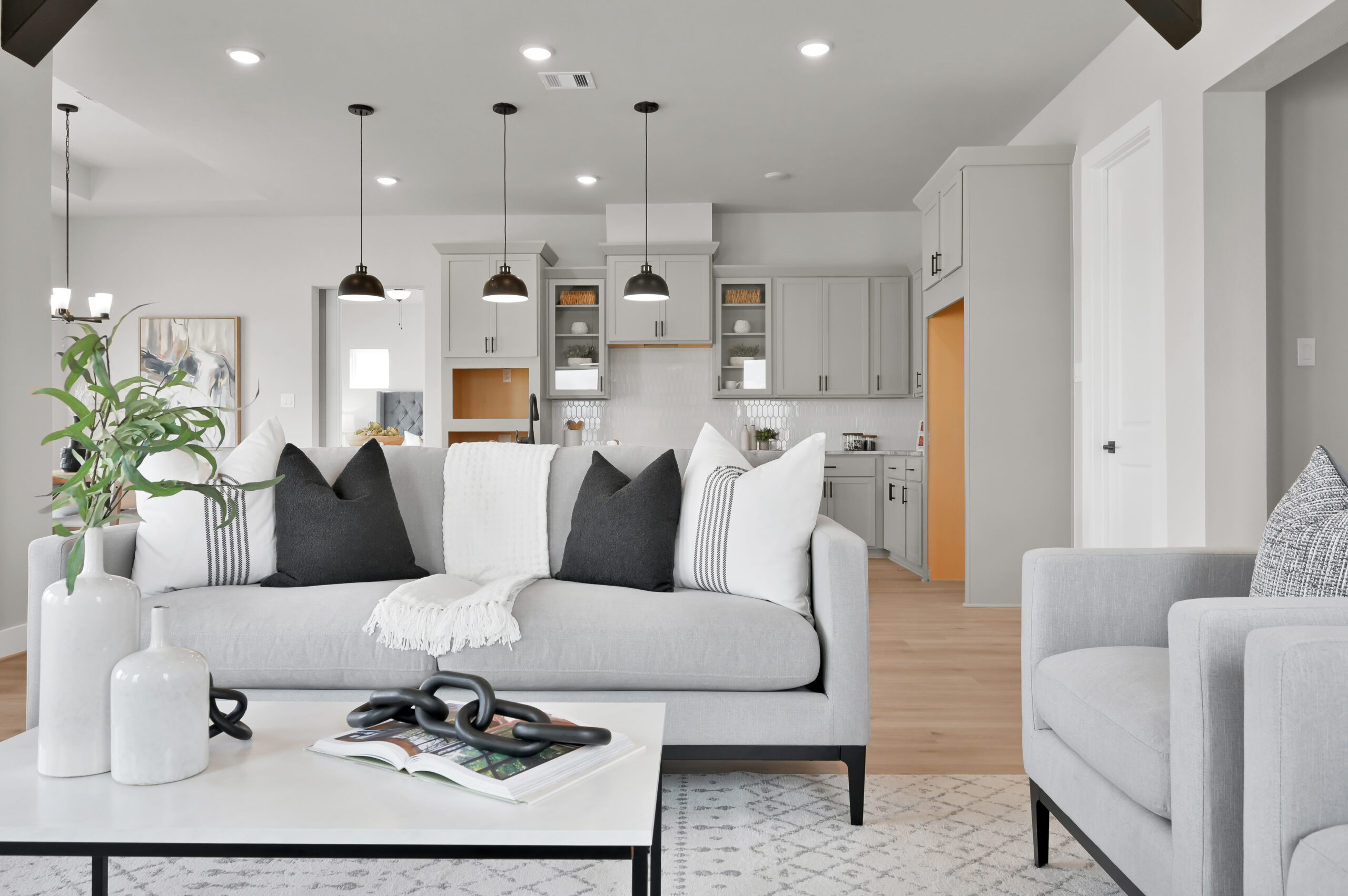 Modern living room with a light gray sofa, black and white pillows, and a white throw blanket. A white coffee table with decorative vases sits on a patterned rug. Open kitchen with pendant lights in the background.
