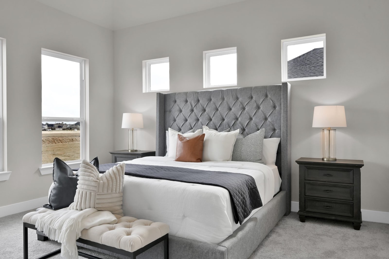 A modern bedroom with a tufted gray headboard, white bedding, and accent pillows. Two dark nightstands with lamps flank the bed. A cushioned bench with a throw blanket sits at the foot of the bed. Four windows let in natural light.