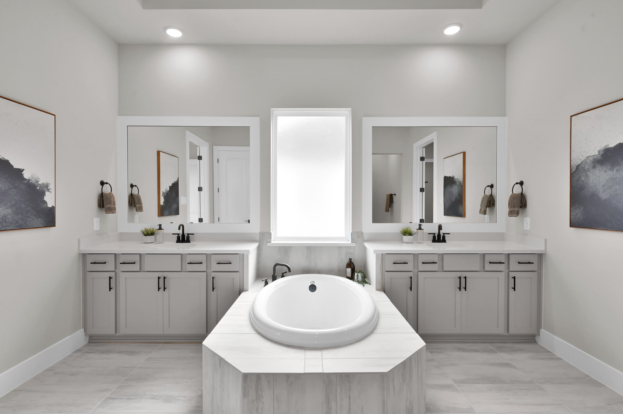 Modern bathroom with a large round bathtub centered between two identical vanities, each with a mirror, sink, and storage cabinets. Soft lighting and minimalist decor create a clean, serene atmosphere.