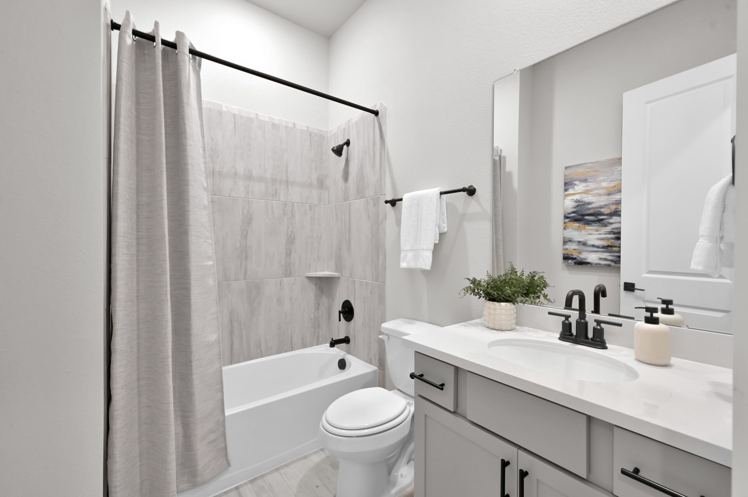 Modern bathroom with light gray walls, a white bathtub with a gray shower curtain, black fixtures, a white toilet, a vanity with a white countertop, a potted plant, and a large mirror above the sink.