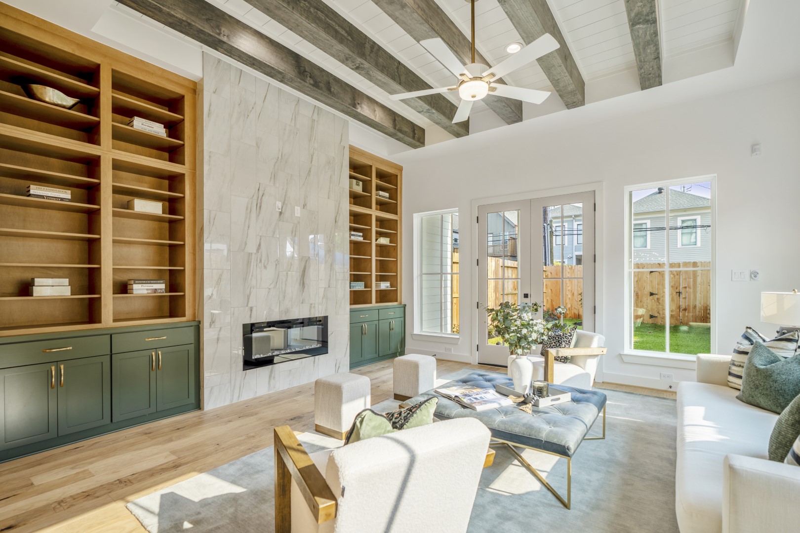 Bright living room with high, beamed ceiling, marble fireplace, built-in wooden shelves with green cabinets, modern furniture, large windows, and glass doors opening to a fenced backyard.