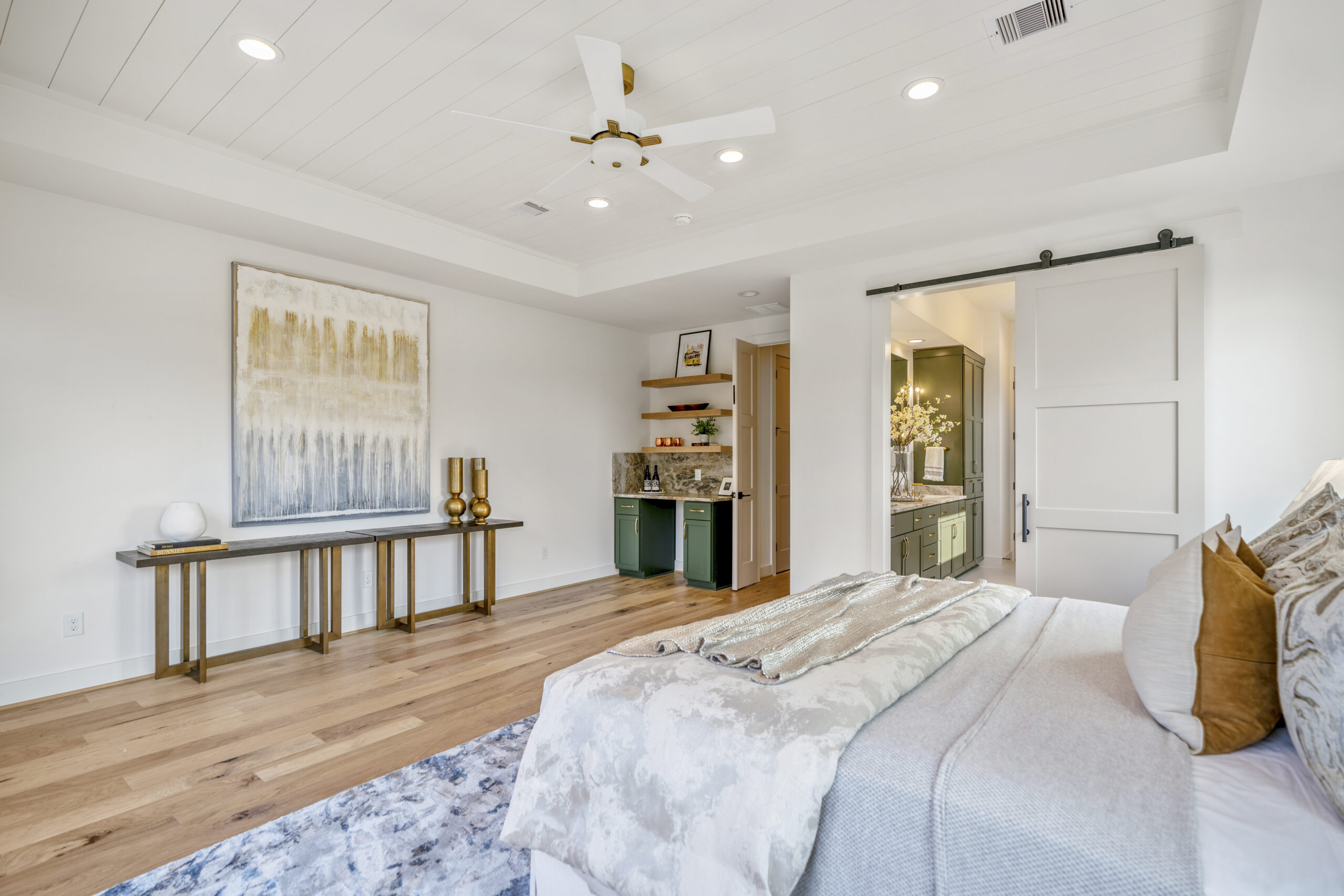 A bright, modern bedroom with a neatly made bed, a large abstract painting above a console table, wood floors, and a sliding barn door opening to a bathroom. Built-in shelves and a small desk area are also visible.