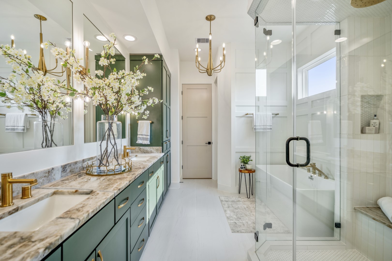 Bright, modern bathroom with green cabinets, marble countertop, gold fixtures, large mirror, glass shower, built-in tub, elegant lighting, and a vase of white flowers on the vanity.