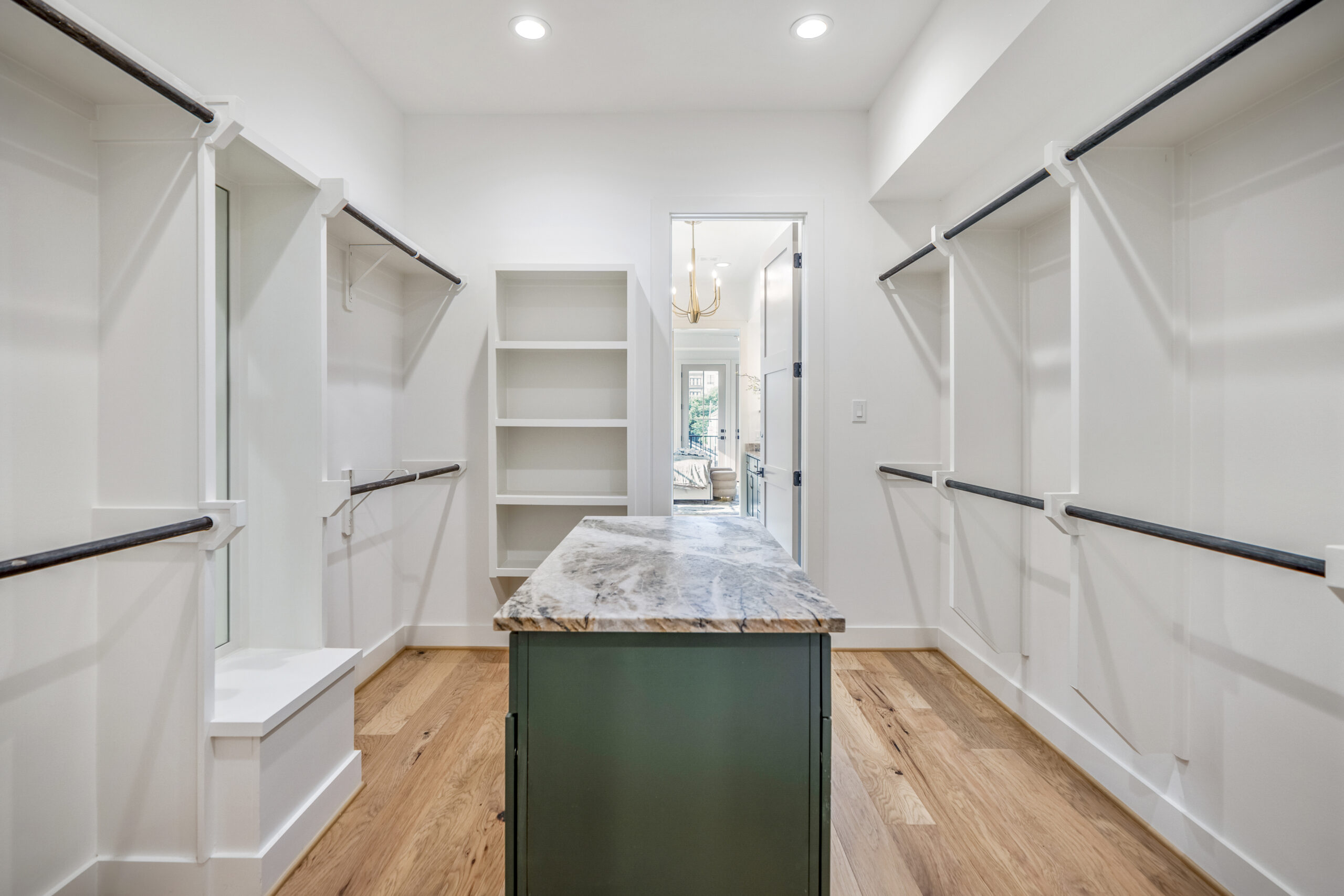 Spacious walk-in closet with white walls, open shelving, multiple hanging rods, wood floor, and a green island with a marble countertop; doorway leads to a well-lit adjoining room.