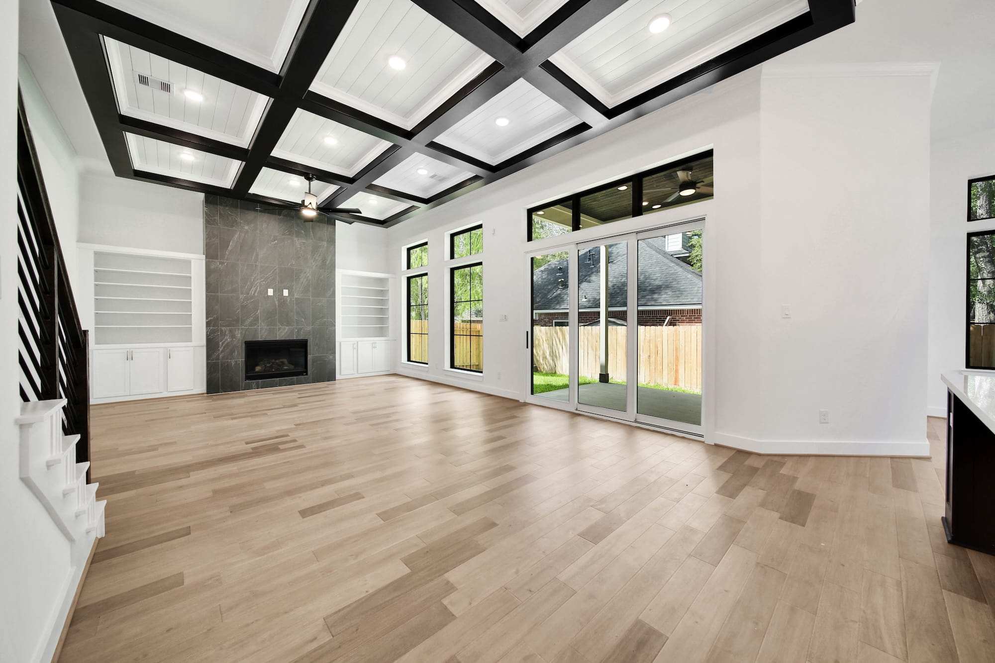 Spacious modern living room with light wood floors, black and white coffered ceiling, large windows, built-in shelves, a dark fireplace, and sliding glass doors opening to a fenced backyard.