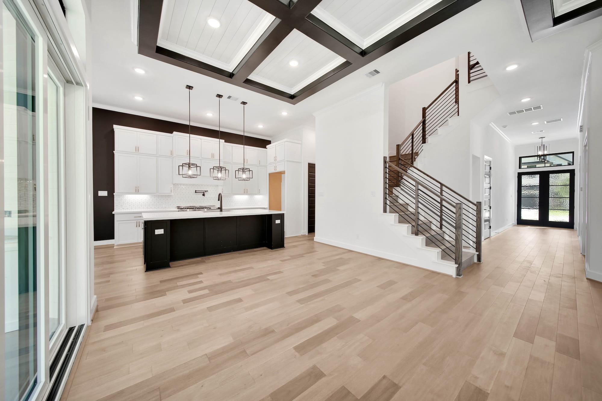 Modern open-concept living space with light wood floors, a black and white kitchen with pendant lights, high coffered ceilings, and a staircase with metal railings near the glass front door.