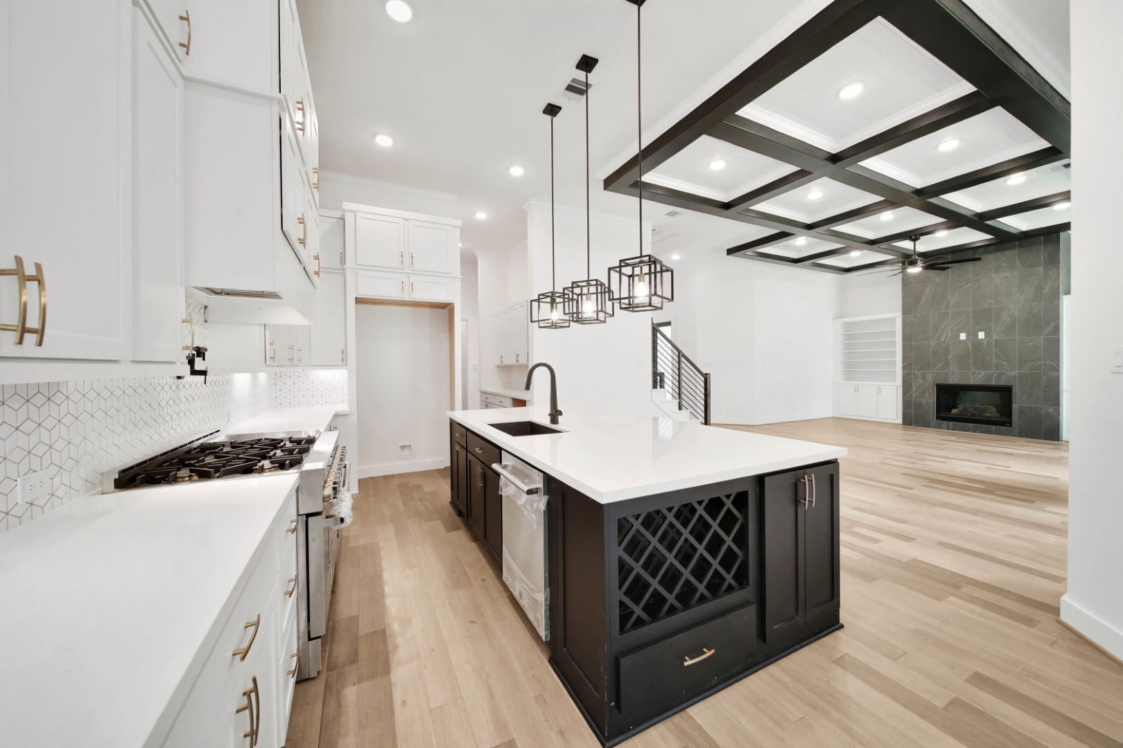 Modern kitchen with white cabinets, black island with wine rack, pendant lights, light wood floors, geometric ceiling design, and open view to living area with fireplace.