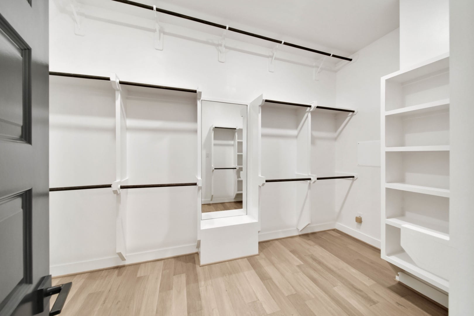Spacious, empty walk-in closet with white shelves, hanging rods, a large mirror in the center, and light wood flooring. The walls and shelving are bright white, creating a clean, modern look.