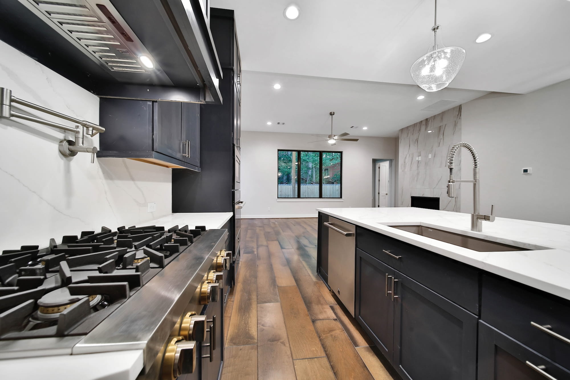 Modern kitchen with black cabinets, stainless steel gas stove, marble countertops, and a large island with a gooseneck faucet. Open layout leads to a spacious living area with wood flooring and large windows.