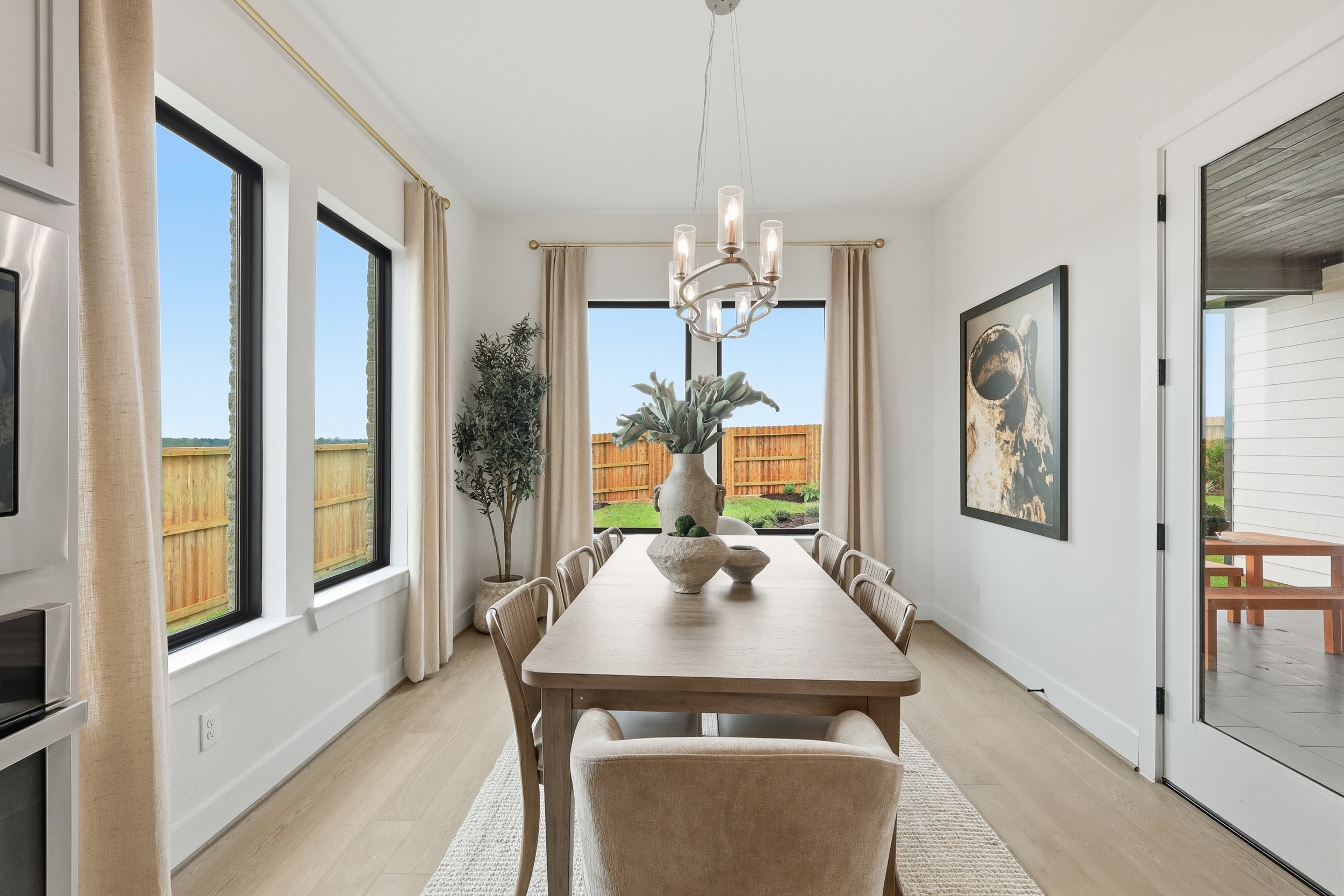 A modern dining room with a wooden table, six beige chairs, a large plant centerpiece, and a contemporary chandelier. Large windows provide natural light and views of a fenced backyard.