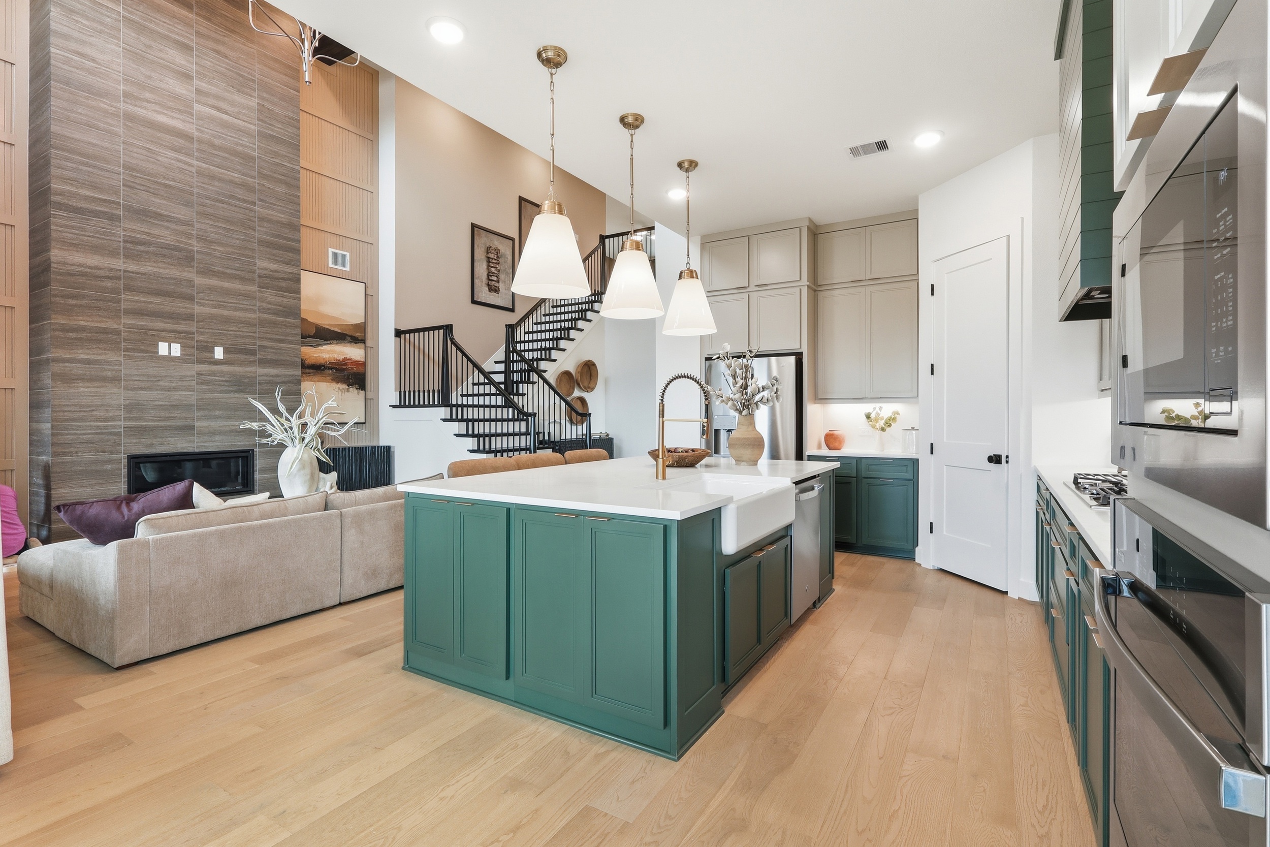Modern open-concept kitchen with green cabinets and island, gold light fixtures, a farmhouse sink, and wood flooring; living area with beige sofa and fireplace in the background, staircase visible.