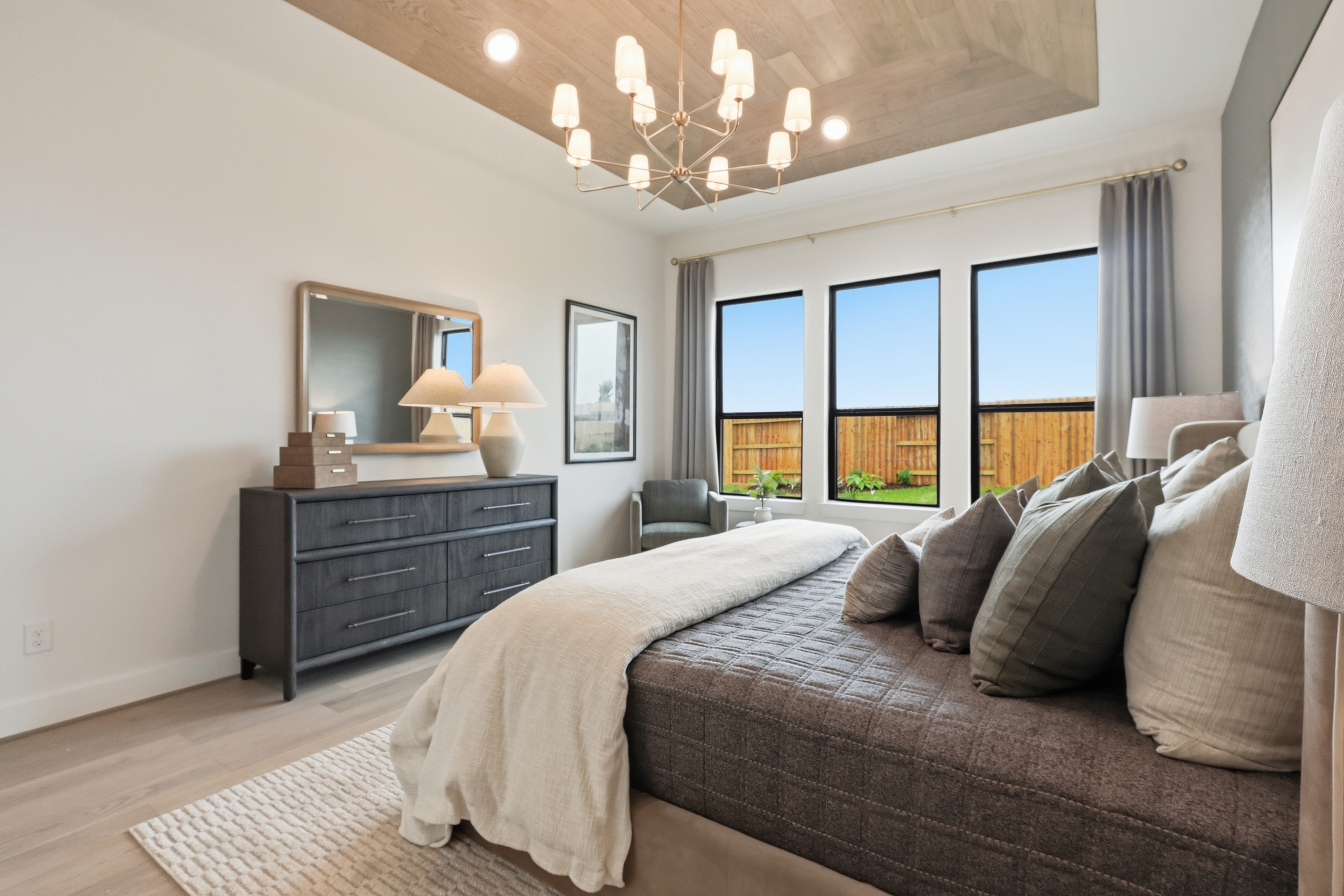 Modern bedroom with a large bed, gray bedding, cushioned pillows, a dresser with a mirror and lamps, an armchair, and three tall windows letting in natural light, overlooking a backyard with a wooden fence.