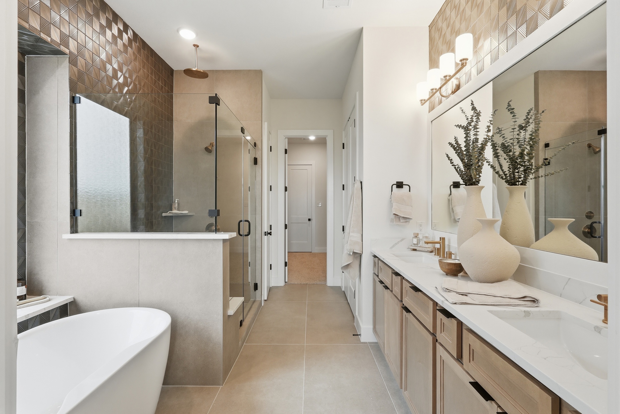 Modern bathroom with a glass-enclosed shower, freestanding bathtub, dual sinks with large mirror, beige cabinets, decorative vases, and geometric wall tiles. Warm lighting and neutral tones create a clean, elegant look.